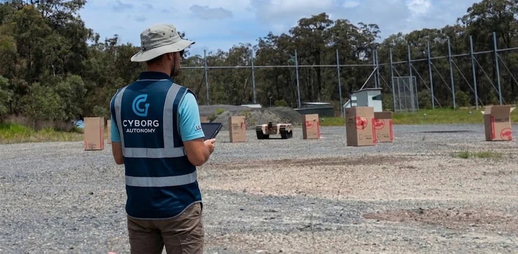 A person wearing a hat and a vest labeled 'CYBORG AUTONOMY' is standing outdoors, looking at a tablet in their hands, with cardboard boxes placed on the ground in front of a chain-link fence.
