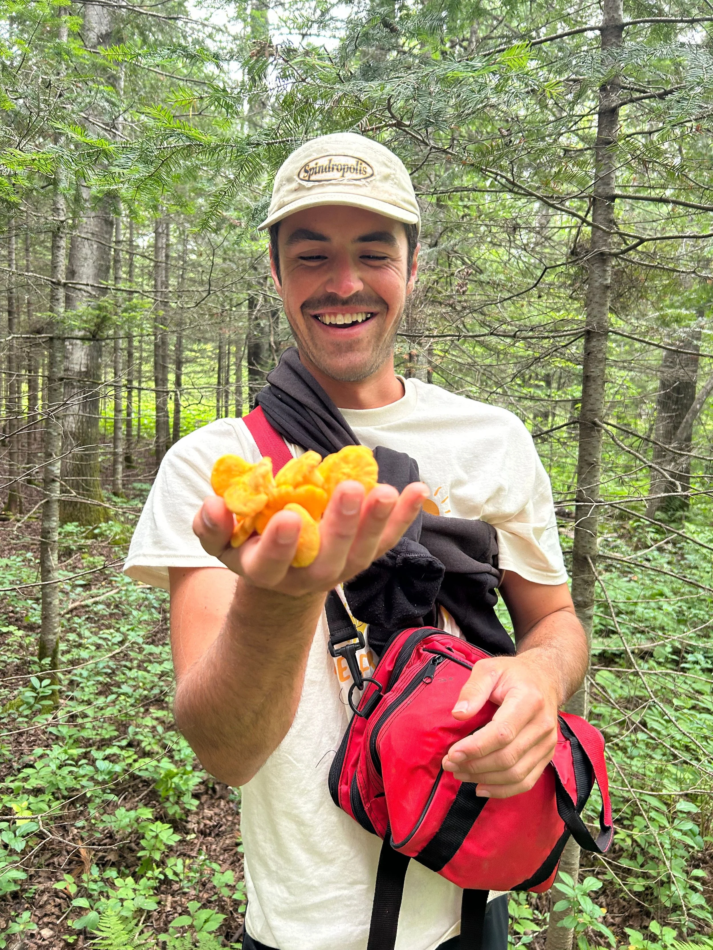 A man smiling in a forest holding yellow puffball fungi in his hand, wearing a beige cap, a white T-shirt, and a red backpack.
