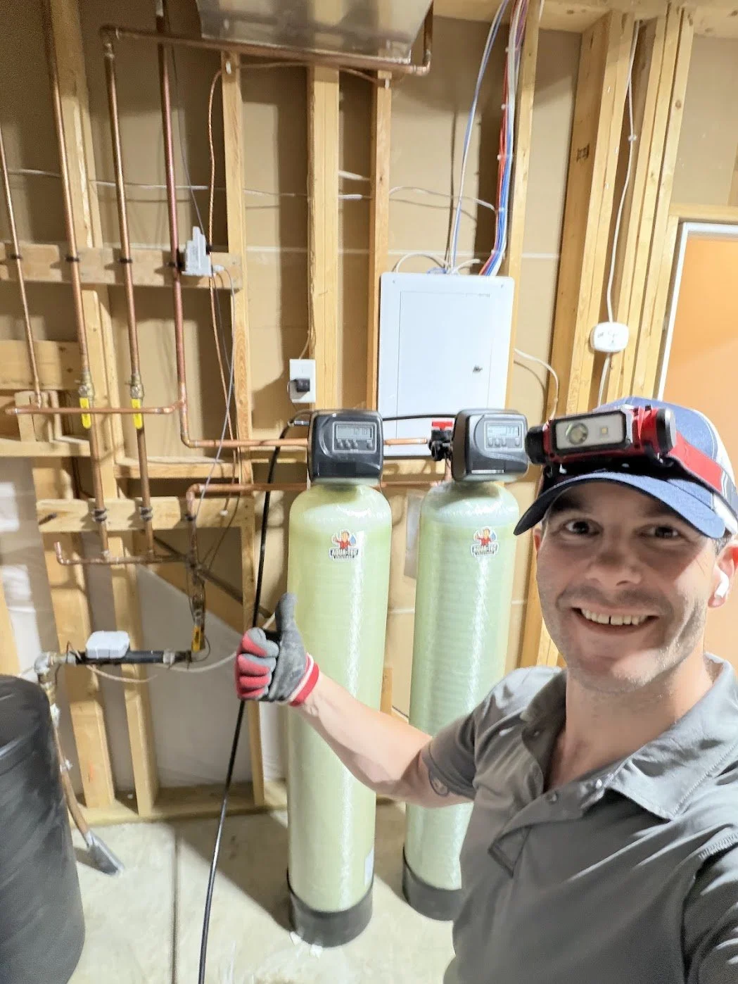 A man with a headlamp and work gloves smiling, giving a thumbs-up, standing in front of water filtration system with two green tanks, exposed wooden framing, and electrical wiring in a home utility room.