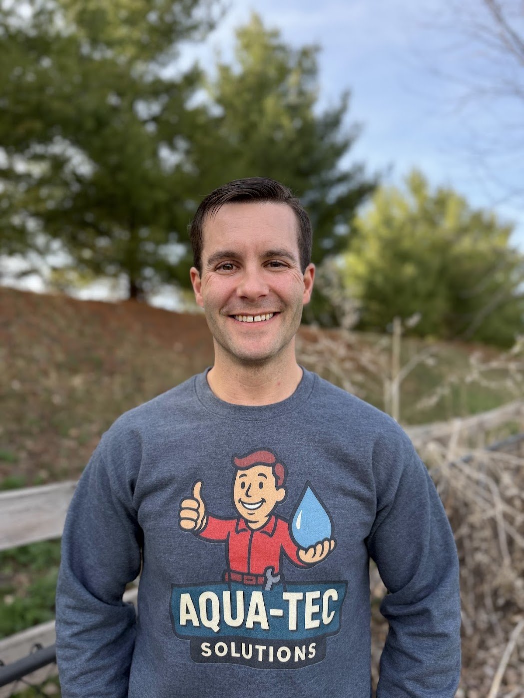A man smiling outdoors wearing a gray sweatshirt with a cartoon character giving a thumbs-up and holding a water droplet, with text reading 'Aqua-Tec Solutions'.