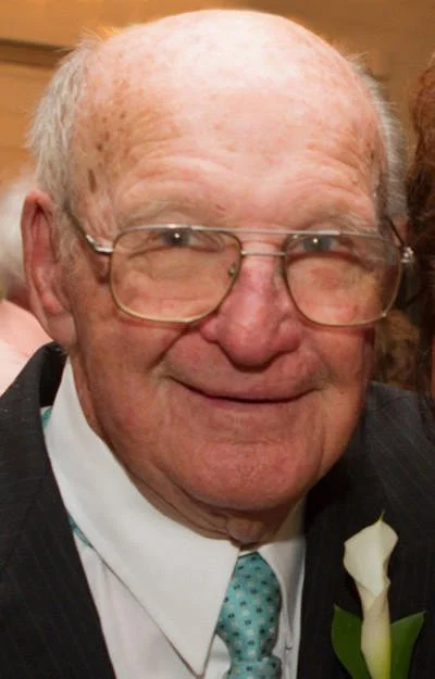 Close-up of an elderly man with glasses, wearing a suit and tie, smiling.