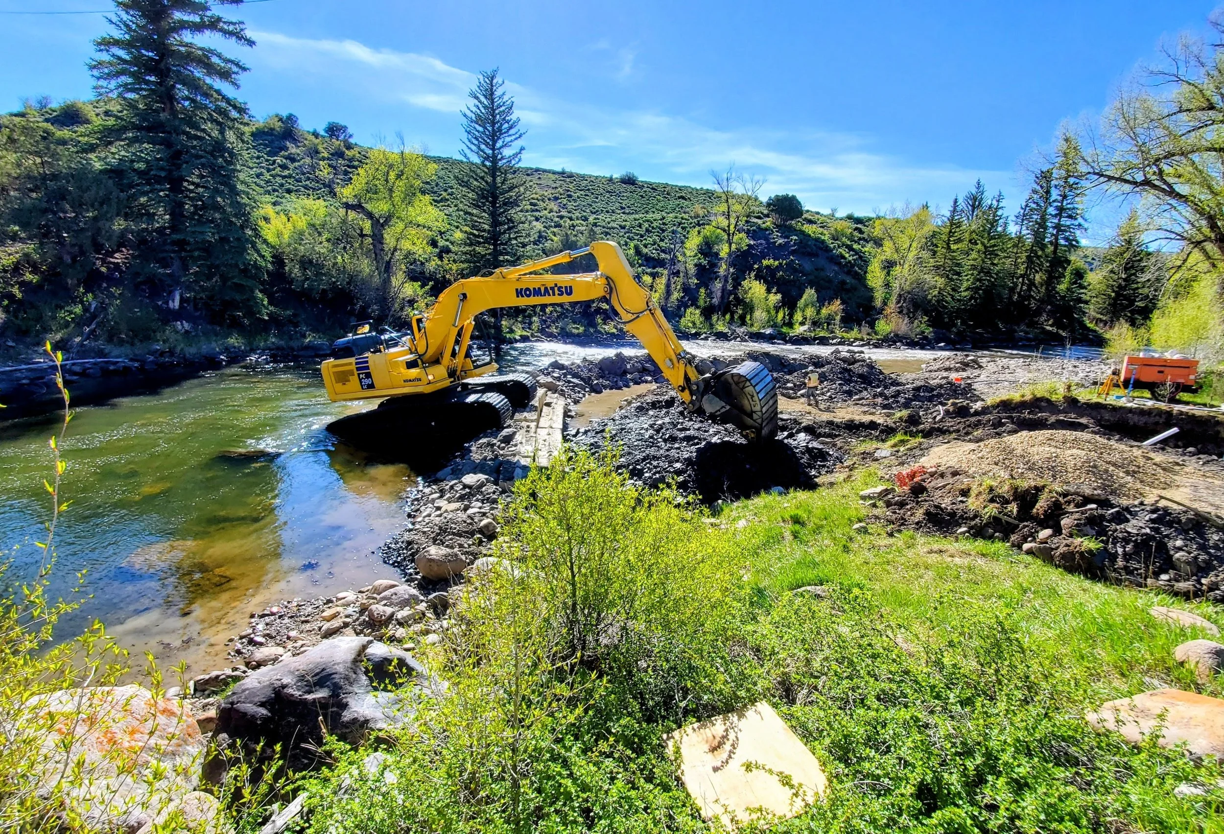 A yellow Komatsu excavator working on a riverbank construction site with water and green trees in the background.
