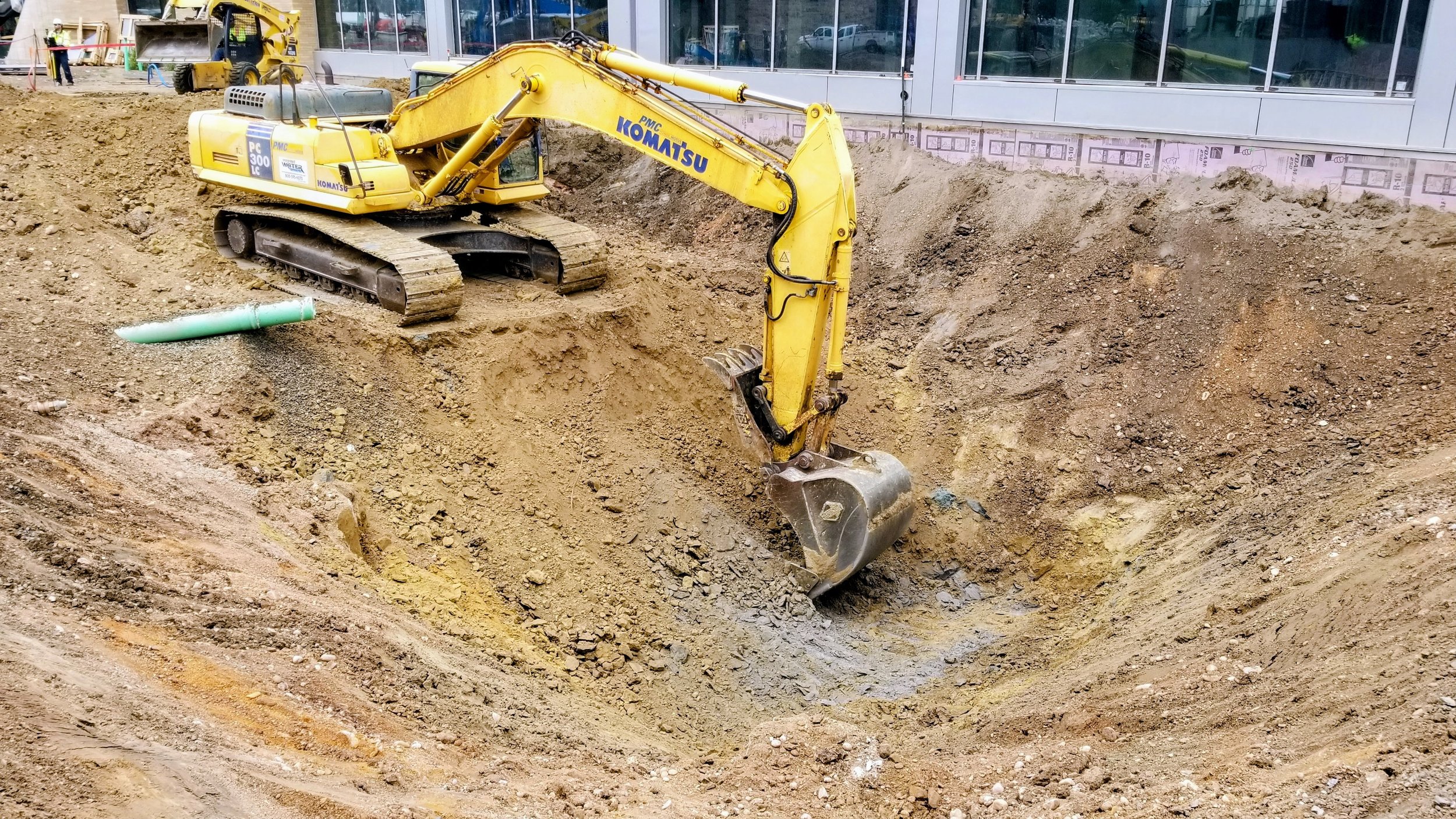 A yellow Komatsu excavator digging a large hole at a construction site next to a glass-front building.