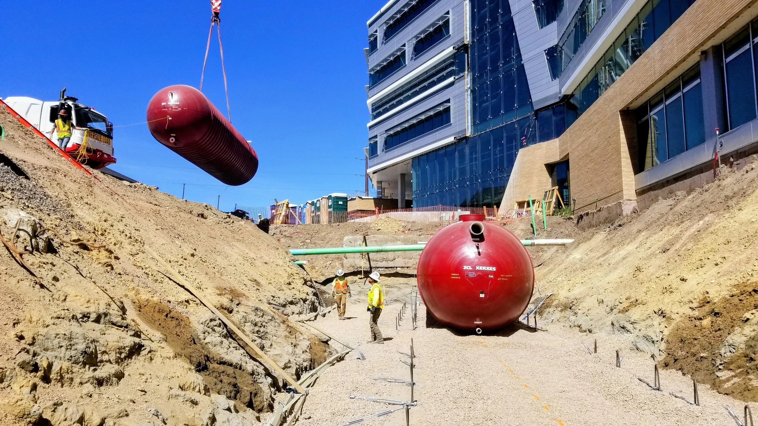 Construction site with two large red spherical tanks, workers wearing safety vests and helmets, modern glass building in background, and clear blue sky.