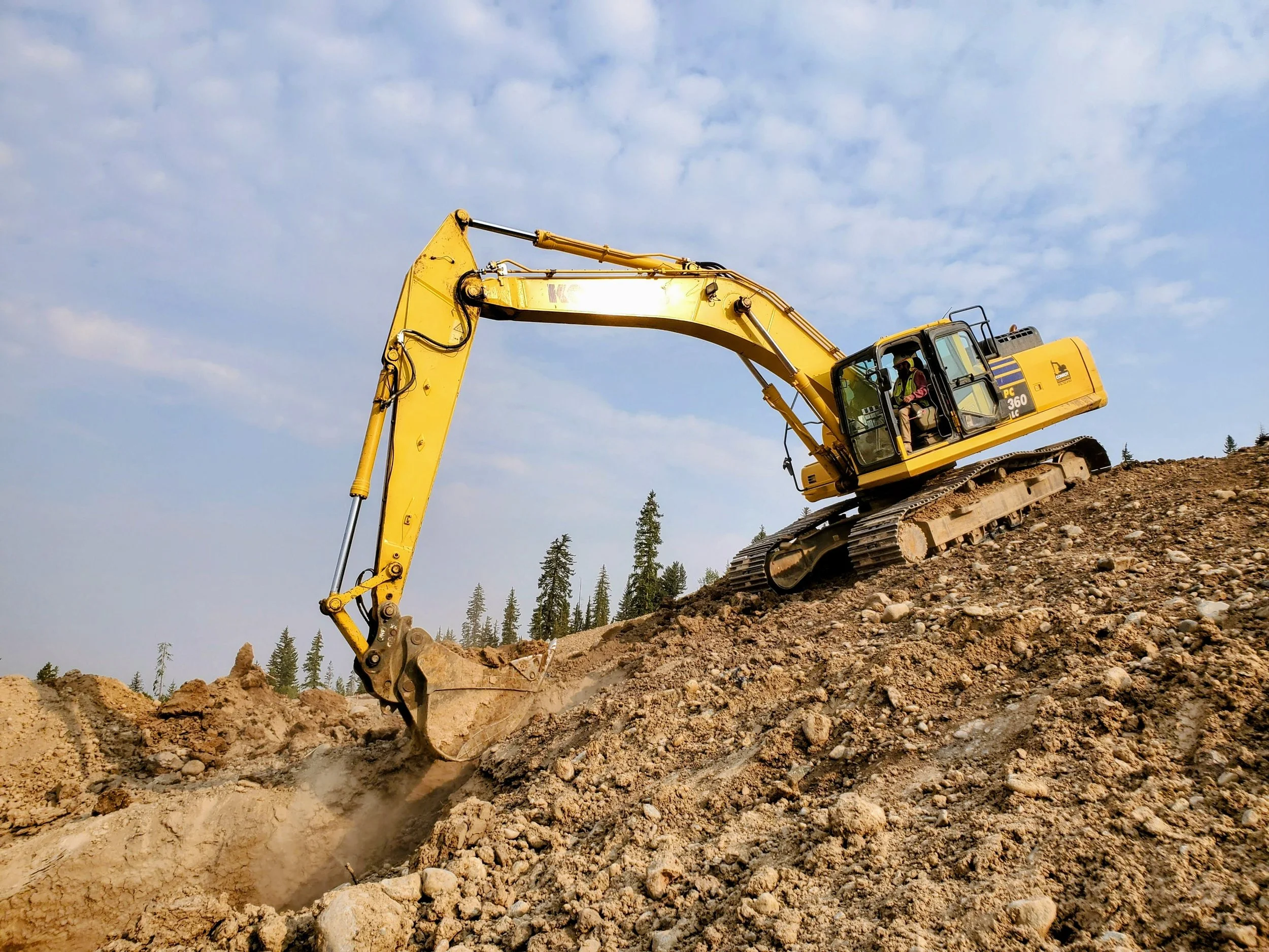 A yellow excavator is digging into a hillside with dirt and rocks, with a partly cloudy sky and trees in the background.