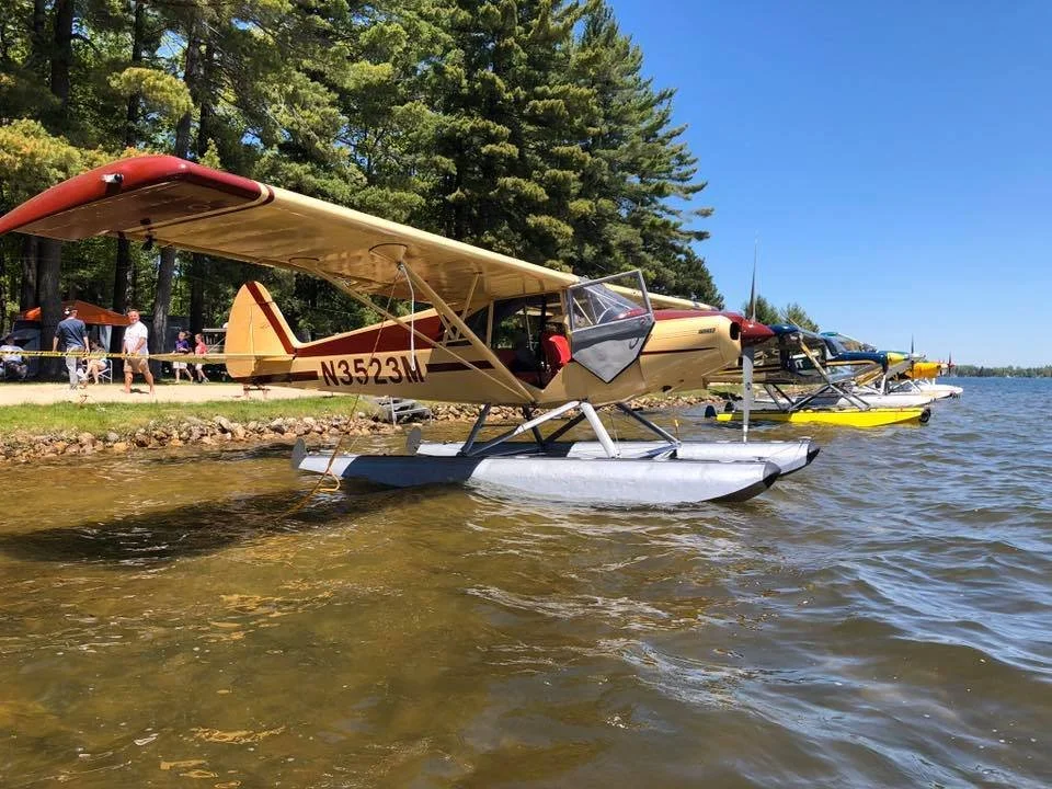 Murphy Rebel and Piper seaplane on floats tied up along a Michigan shoreline on a sunny day.