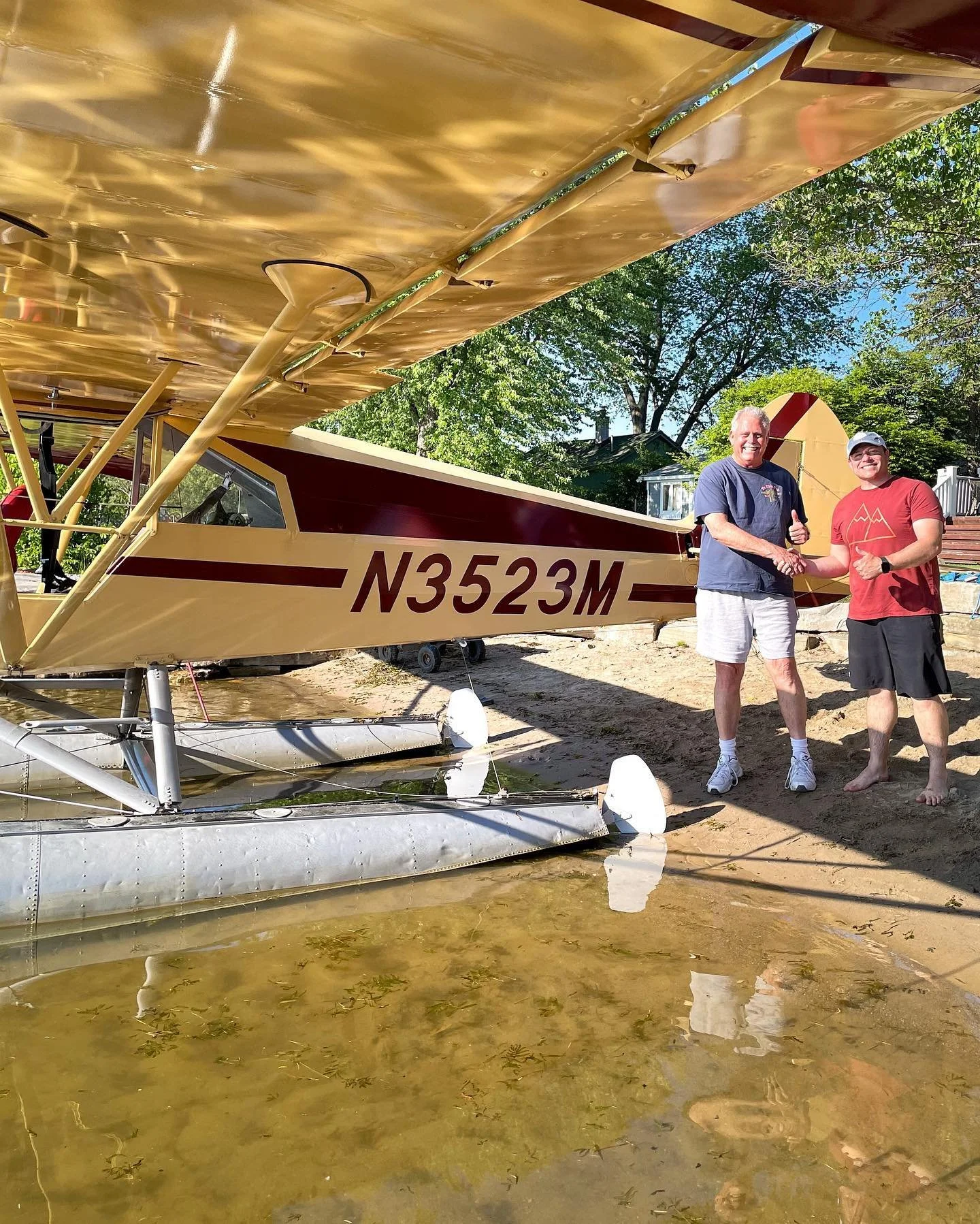 Two people standing beside a seaplane on floats at a sandy lakeshore in Michigan