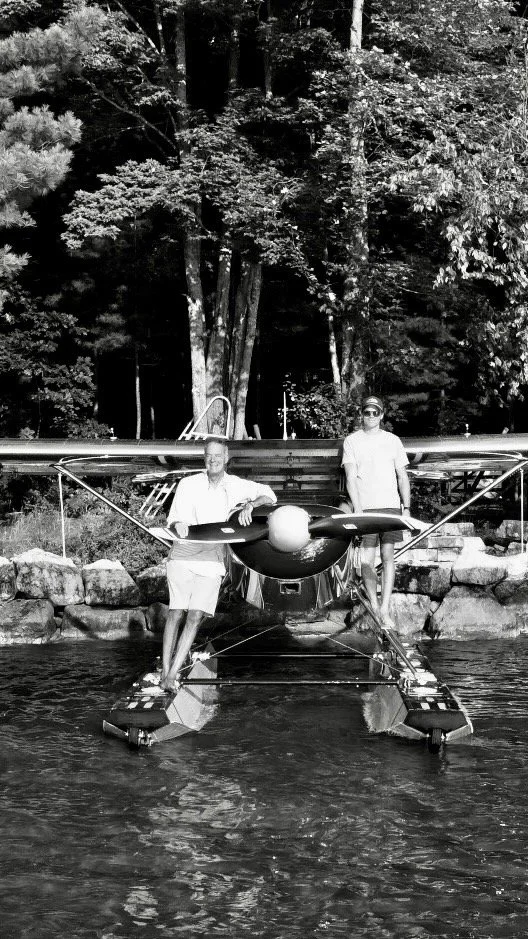 Two pilots standing on the floats of a Murphy Rebel seaplane on a calm Michigan lake, with forested shoreline behind them.