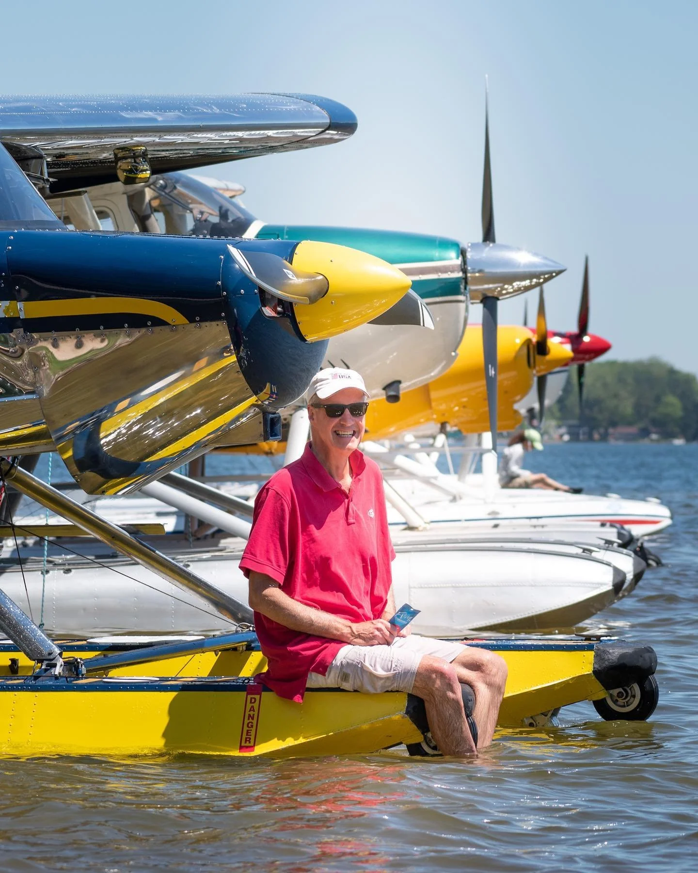 Seaplane pilot seated on floats beside multiple seaplanes on a Michigan lake