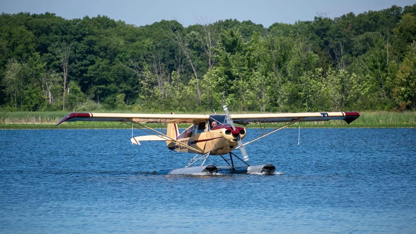 Yellow single-engine seaplane touching down on a calm Michigan lake, surrounded by green shoreline trees.