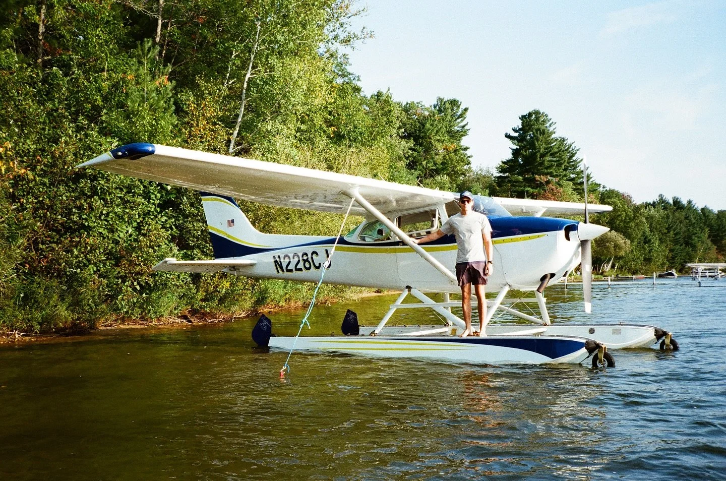ilot standing on the float of a single-engine seaplane moored on a Michigan lake with wooded shoreline.