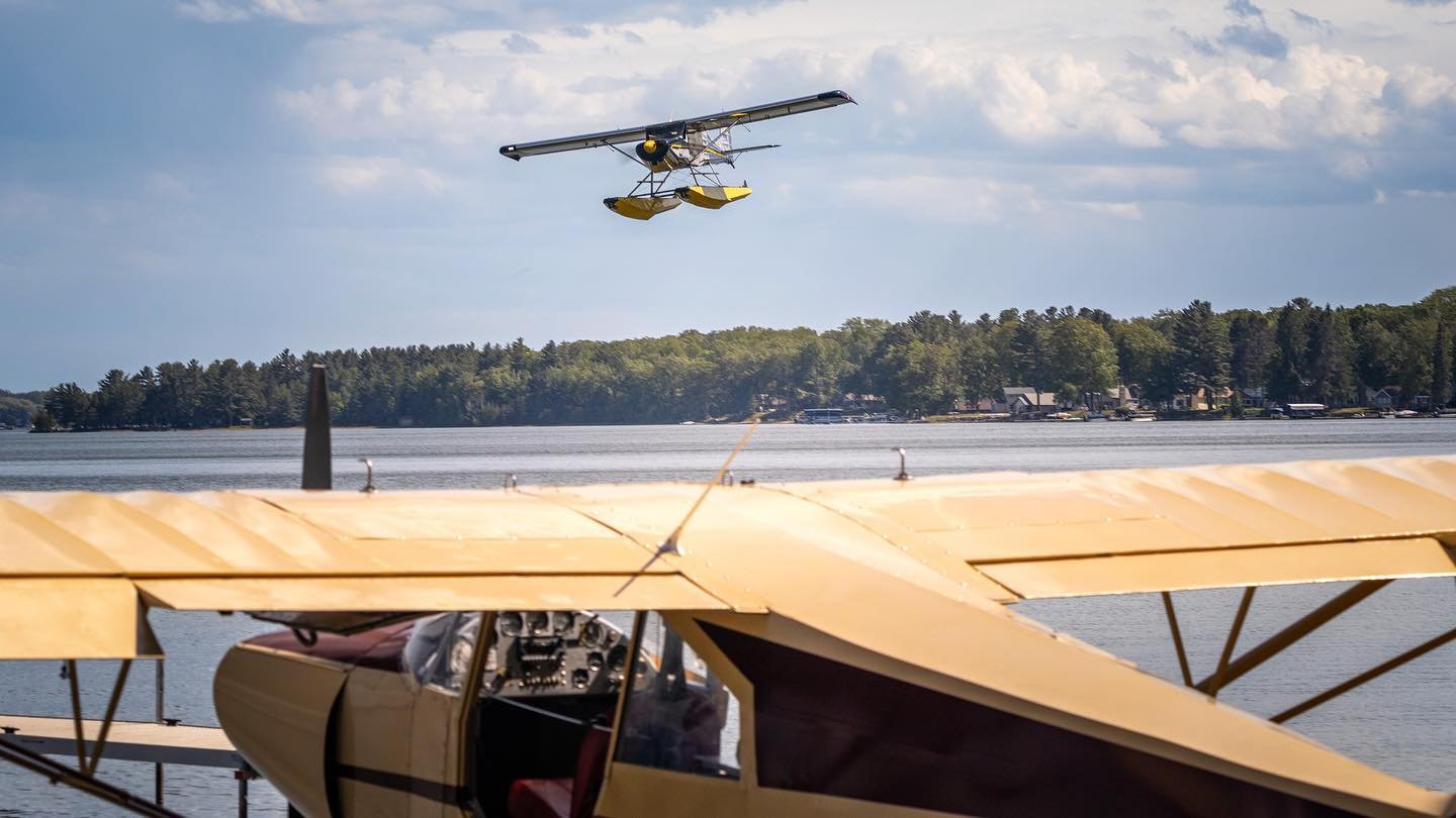 Floatplane airborne over a lake with docked seaplane in the foreground