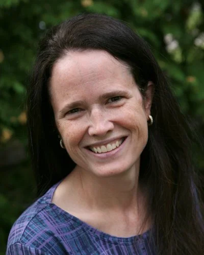Smiling woman with dark hair wearing earrings and a purple patterned shirt outdoors.