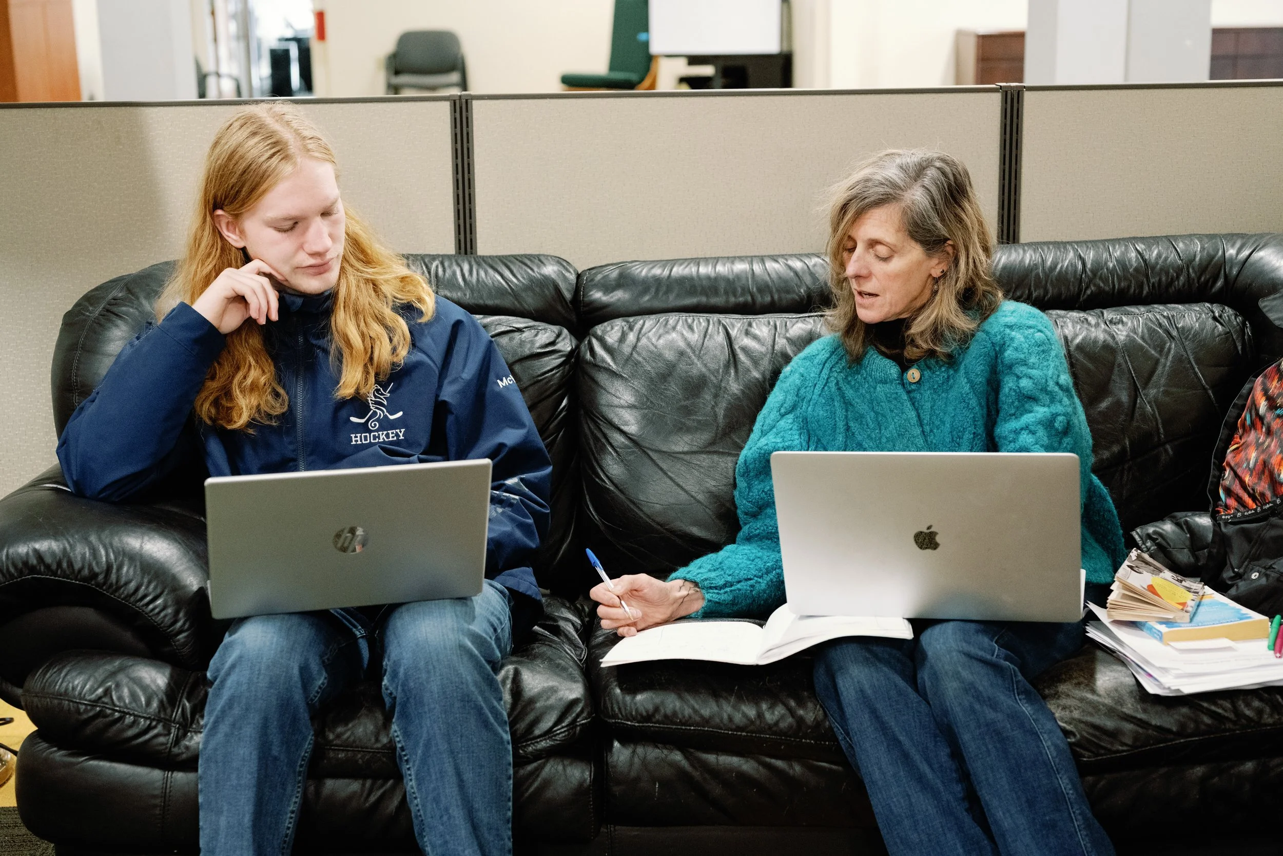 A young woman with long red hair sitting on a black leather couch next to an older woman with gray hair. Both have laptops on their laps and are engaged in conversation, with notebooks and papers nearby.