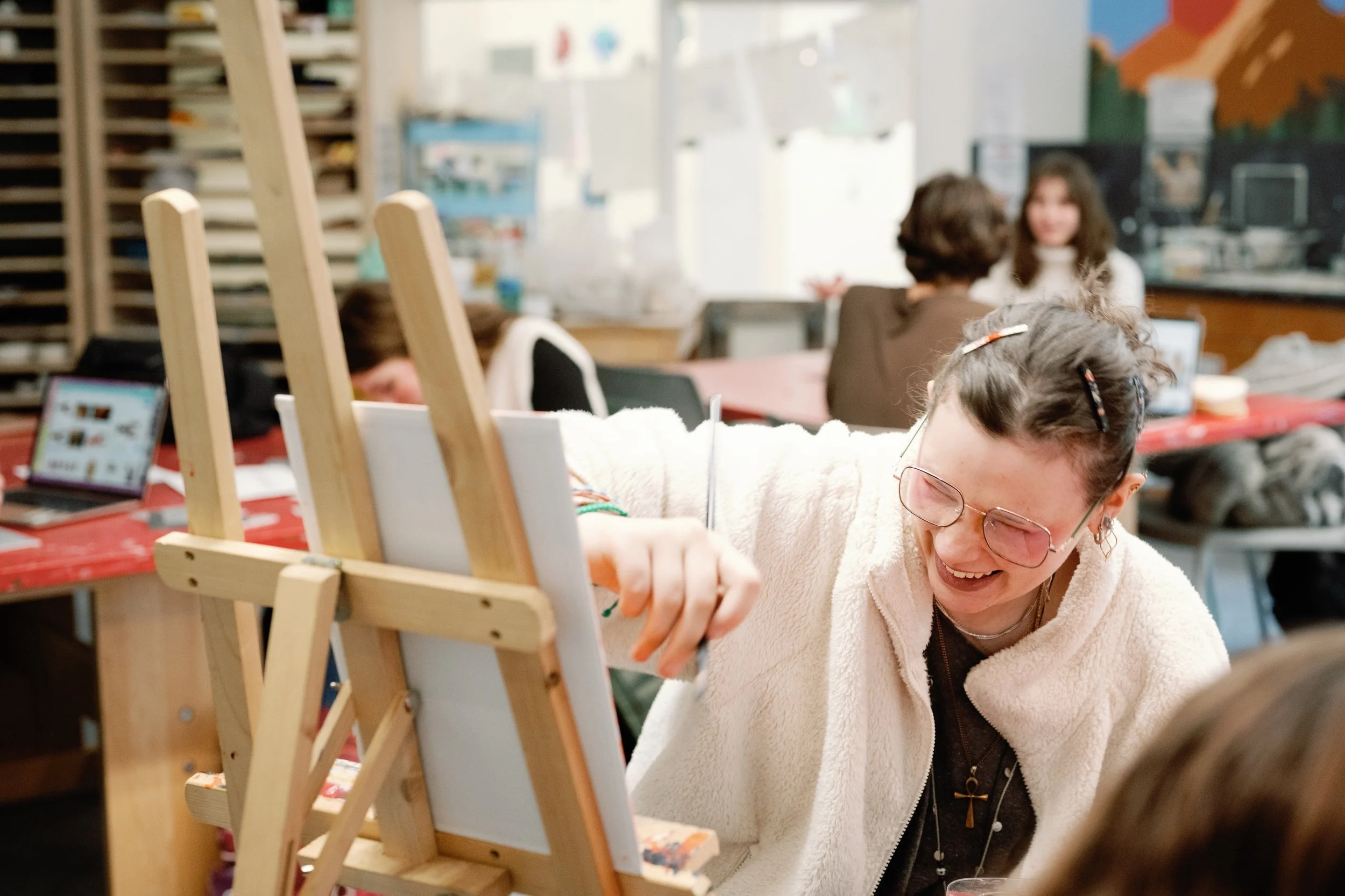 A young woman with glasses and a fleece jacket laughing as she paints on a canvas in an art classroom with other students and teachers in the background.