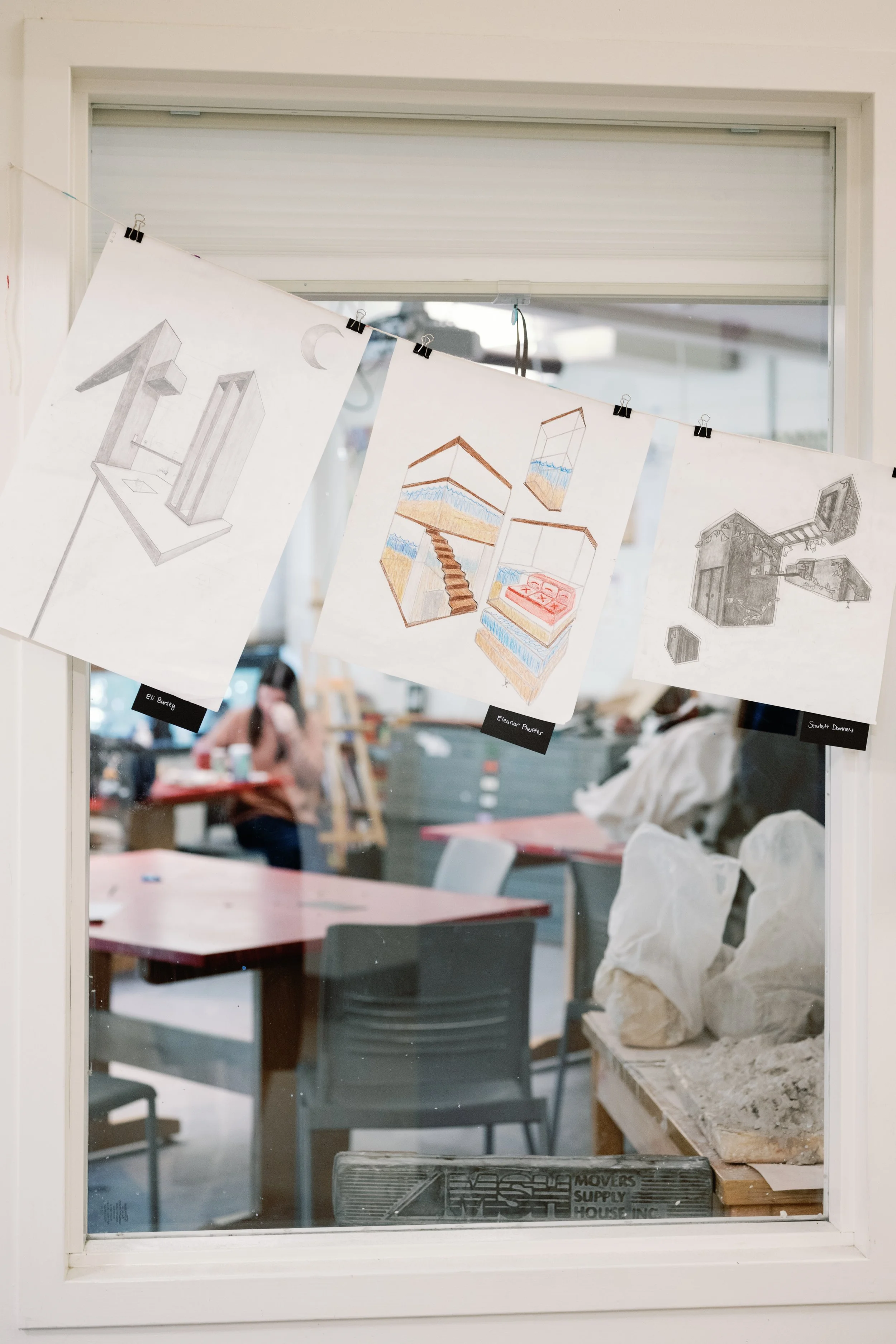 Sketches and drawings of architectural models hanging on a string in a room, with furniture and a person in the background visible through the window.