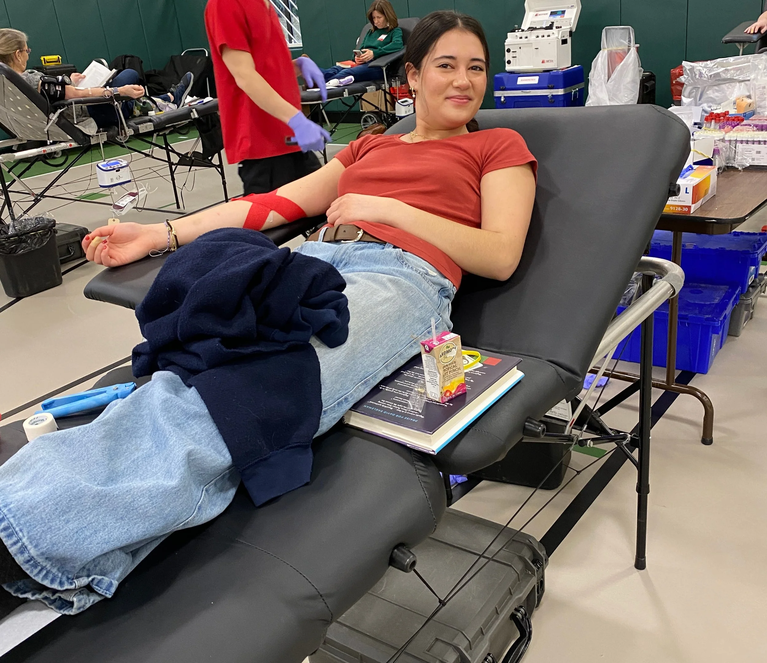 A young woman with dark hair, wearing a red t-shirt and jeans, is lying on a medical chair donating blood. She is smiling at the camera. A red tourniquet is on her upper arm, with a needle inserted. Medical supplies and other donors are visible in the background.