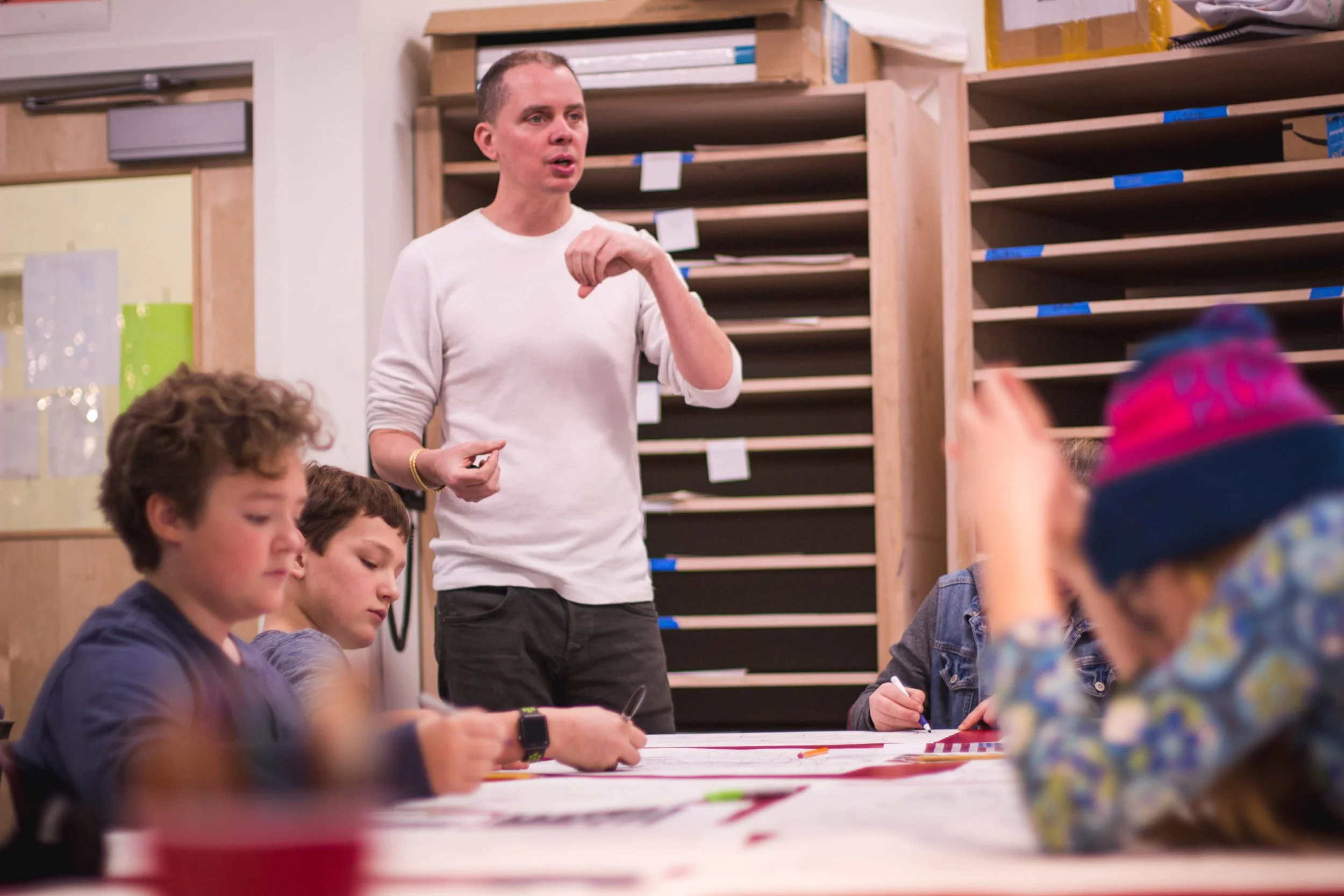 A man standing and speaking to a group of children sitting at a table in a classroom or art room, with shelves behind him filled with papers and supplies.