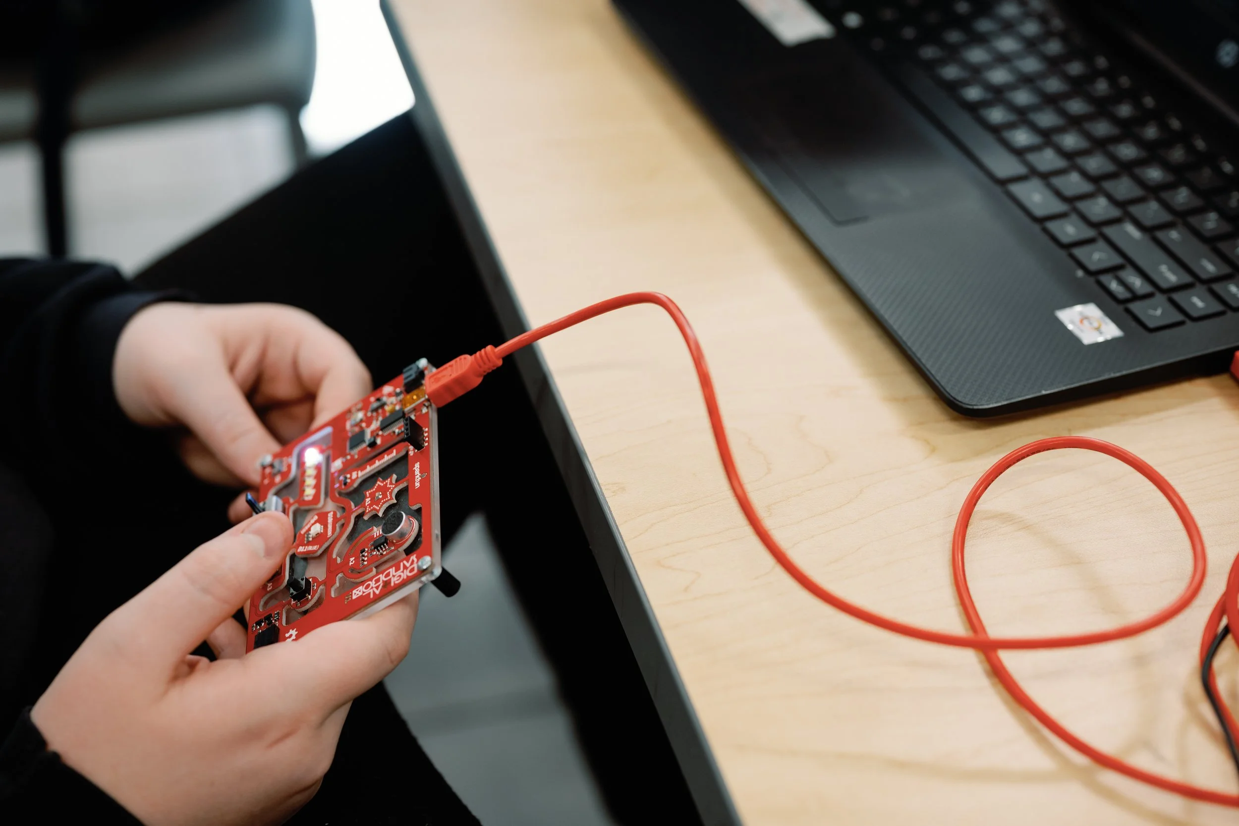 Person holding a red circuit board connected to a laptop on a wooden desk.