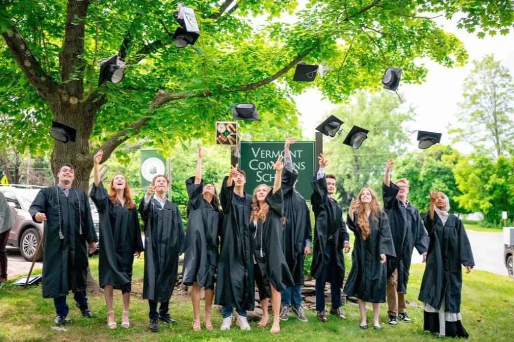 Group of graduates in caps and gowns celebrating outdoors, throwing their caps into the air near a sign that reads 'VERMONT COLLEGES SCHOOL' with lush green trees in the background.