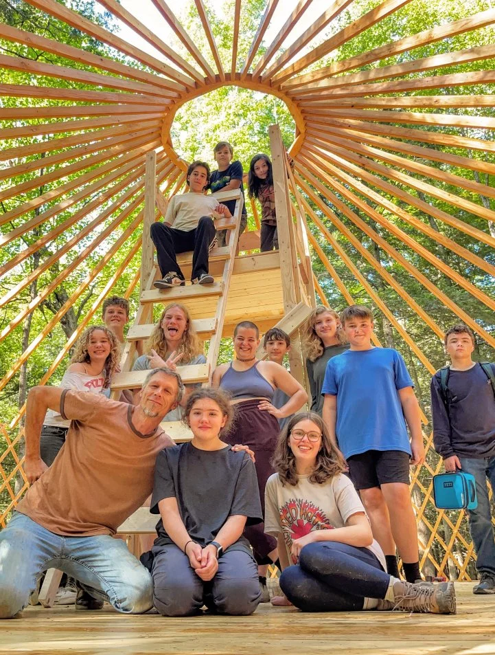 Group of people, including children and adults, gathered inside and around a wooden treehouse with a circular opening, surrounded by green trees in a forest setting.