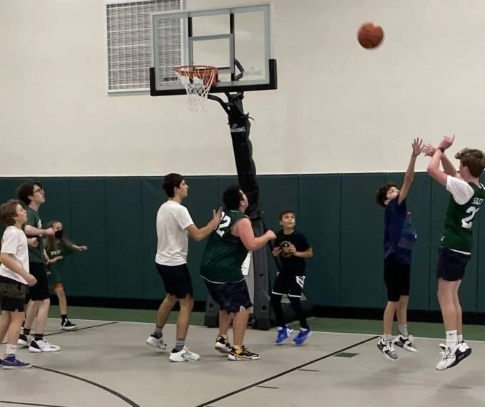 Kids playing basketball in indoor gym, some jumping for shot near the hoop, others watching or waiting.