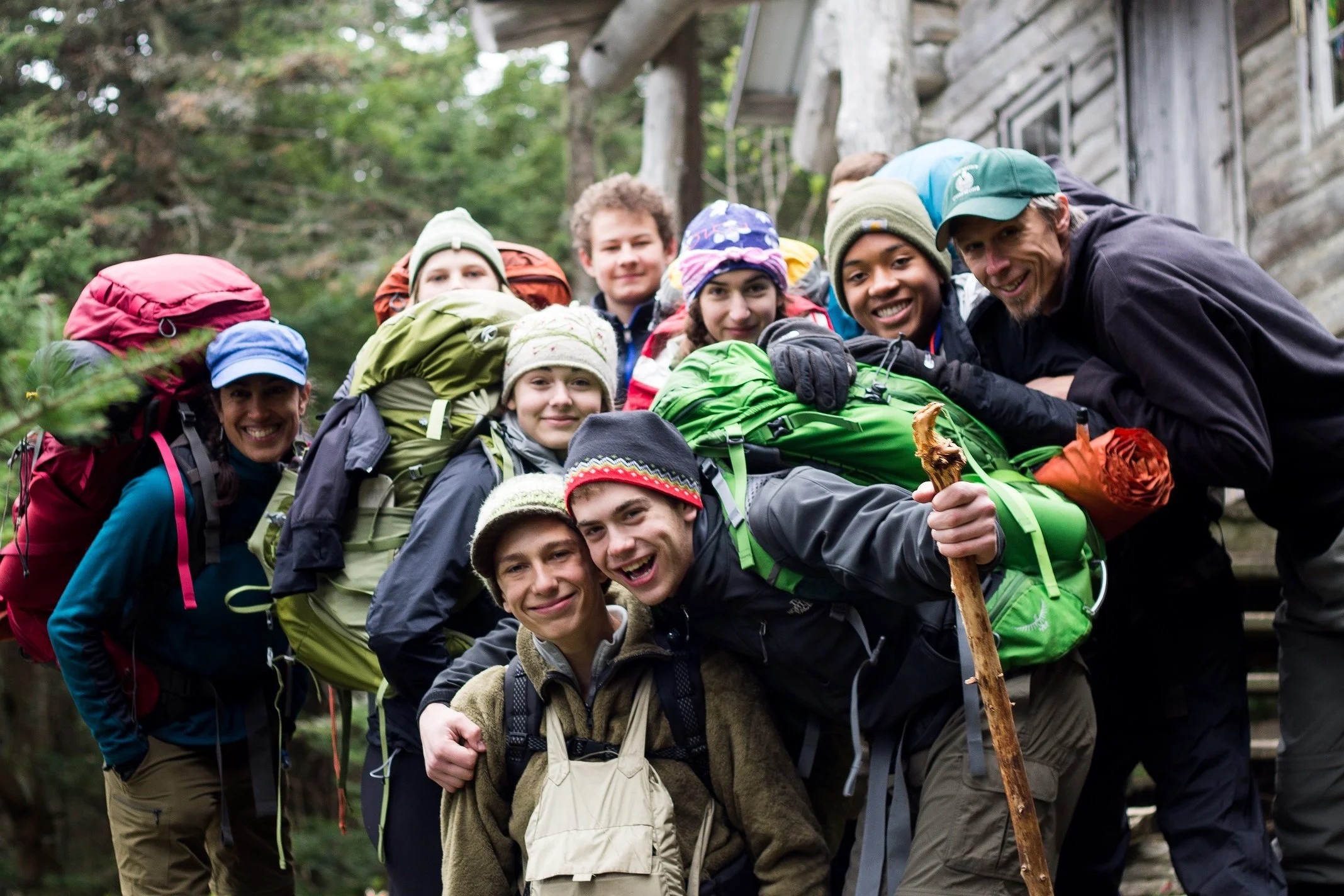 Group of people with backpacks hiking outdoors in a forest near a cabin, smiling and posing for the camera.