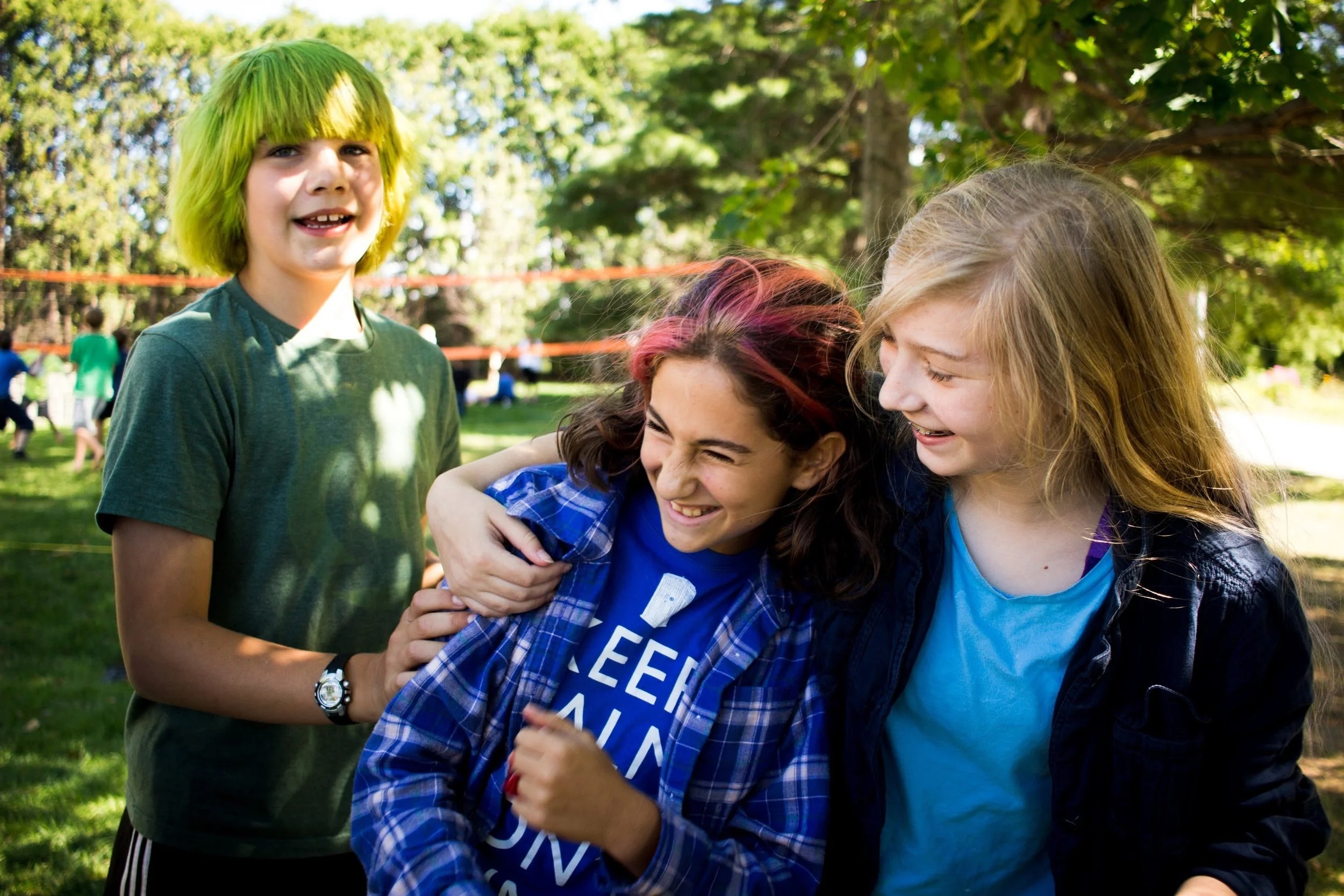 Three young students laughing and hugging outdoors in a sunny park with trees and other children in the background.