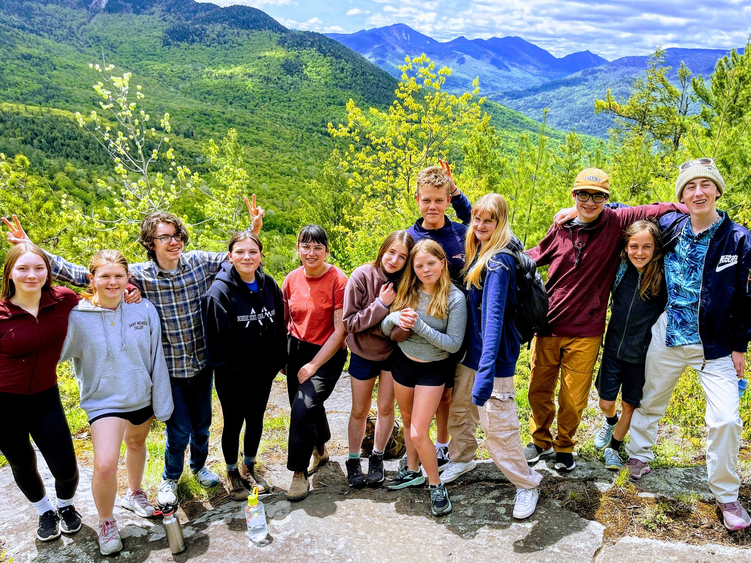 Group of children and teenagers hiking and posing together outdoors with lush green mountains and trees in the background.