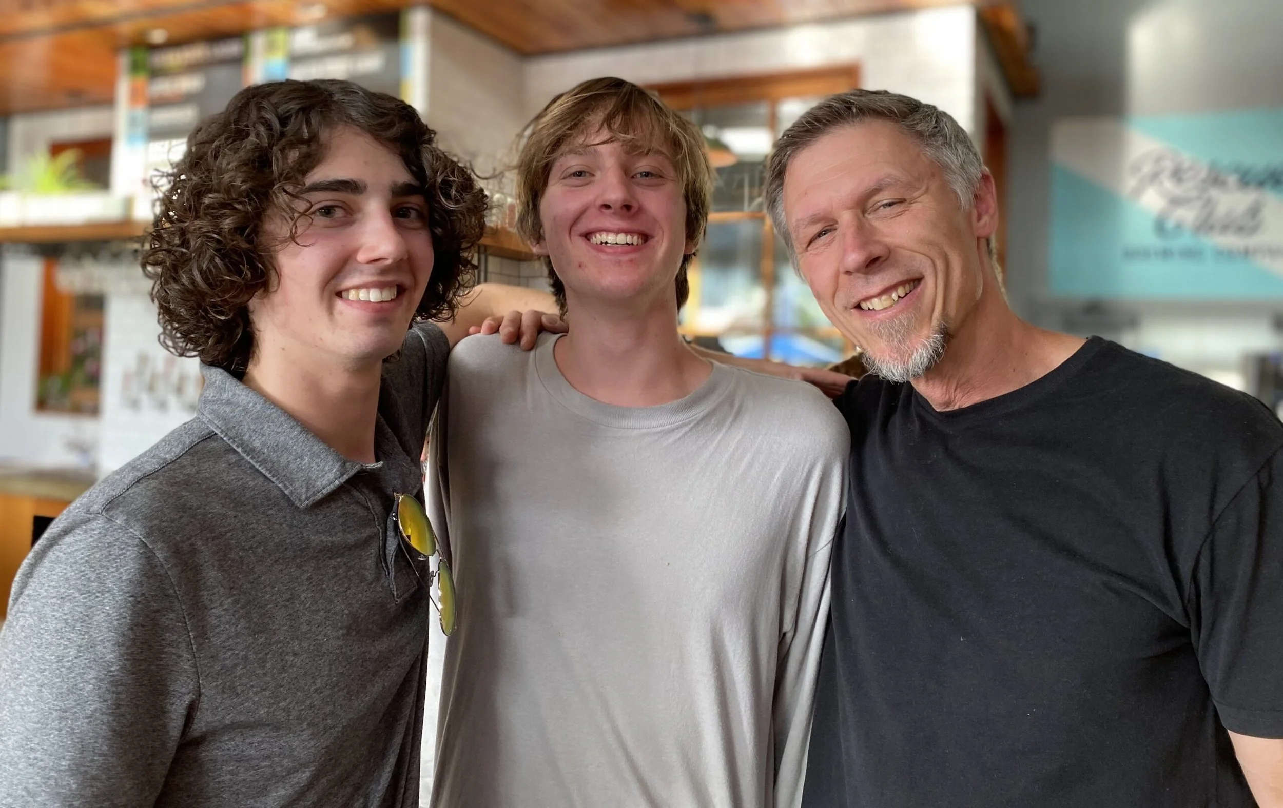 Three smiling men standing close together in a casual setting, with one man's arm around the other two, in a well-lit indoor space.
