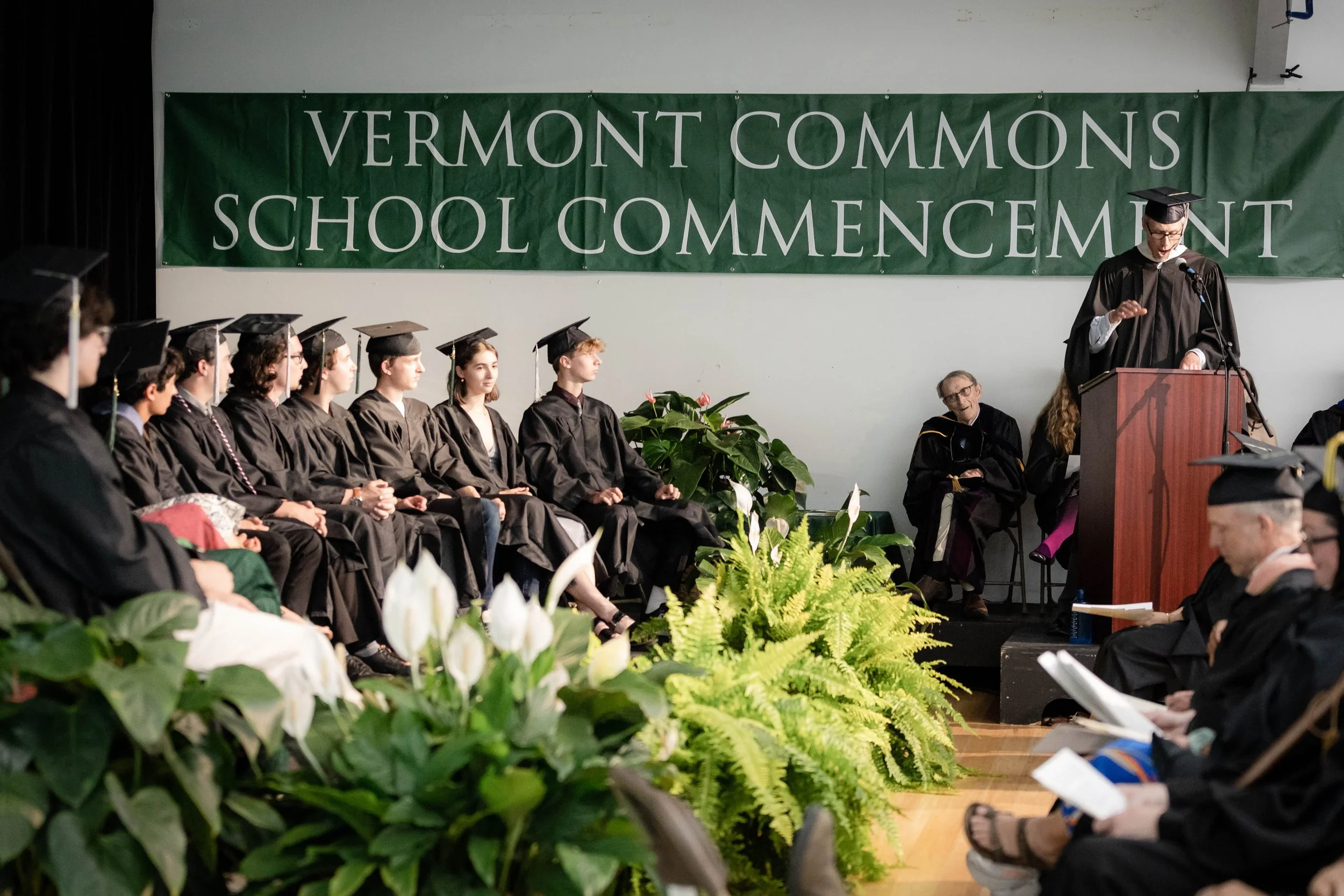 A graduation ceremony at Vermont Commons School where graduates in black caps and gowns are seated on stage, listening to a speaker at a podium. There are large green plants and a banner reading "Vermont Commons School Commencement" in the background.