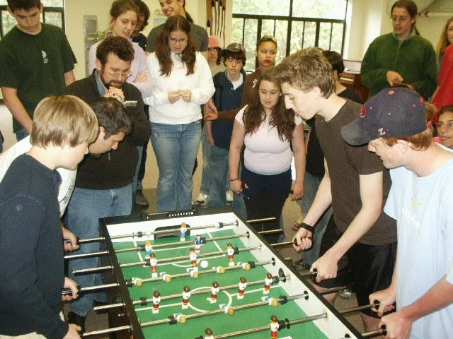 Children and adults playing or watching a game of foosball in an indoor setting with large windows.