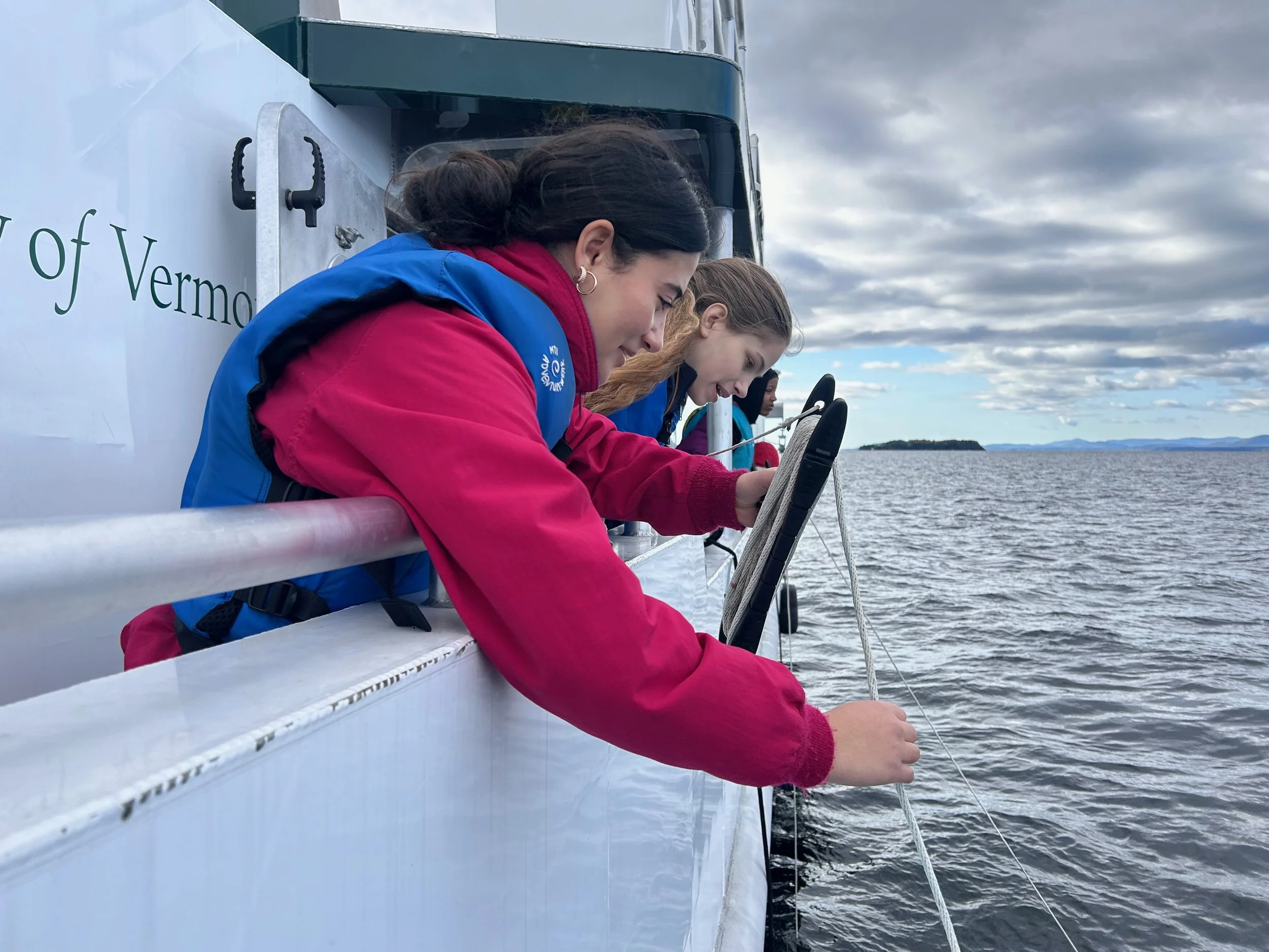 Three girls leaning over the side of a boat, looking at catch or device in the water during cloudy weather.