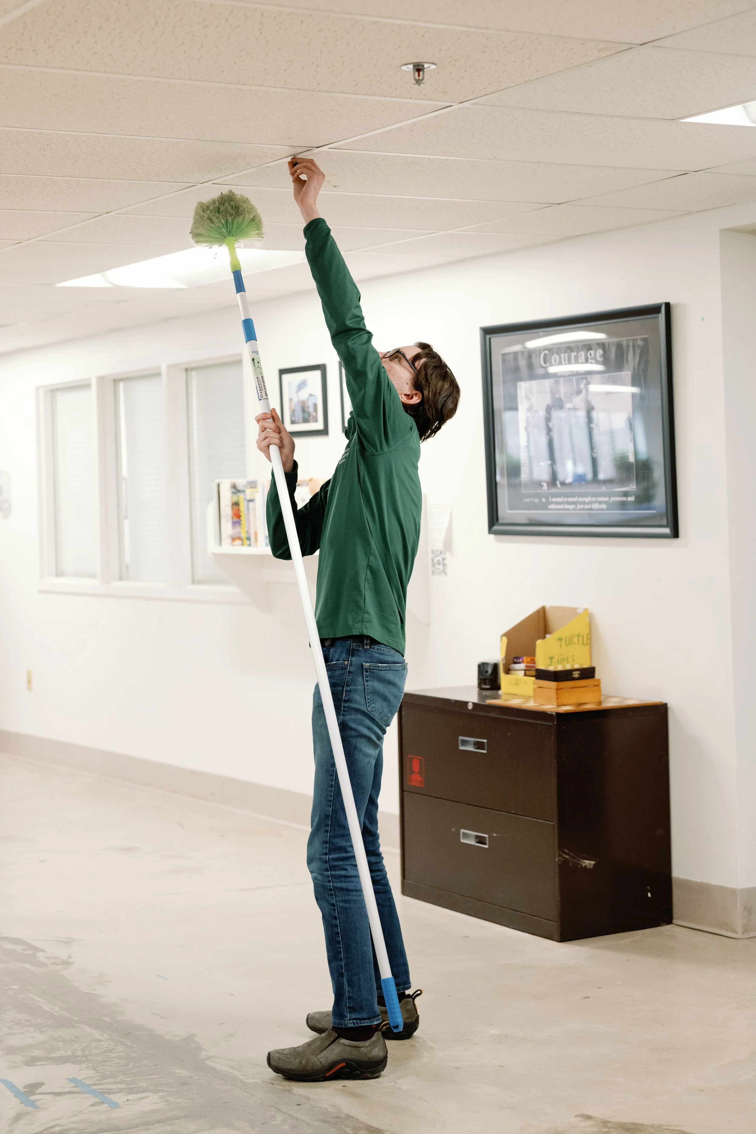 A person using a duster with an extended handle to clean the ceiling of an indoor space.