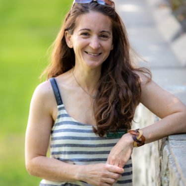 Woman with long brown hair smiling, wearing a striped sleeveless top, leaning on a white fence outdoors.