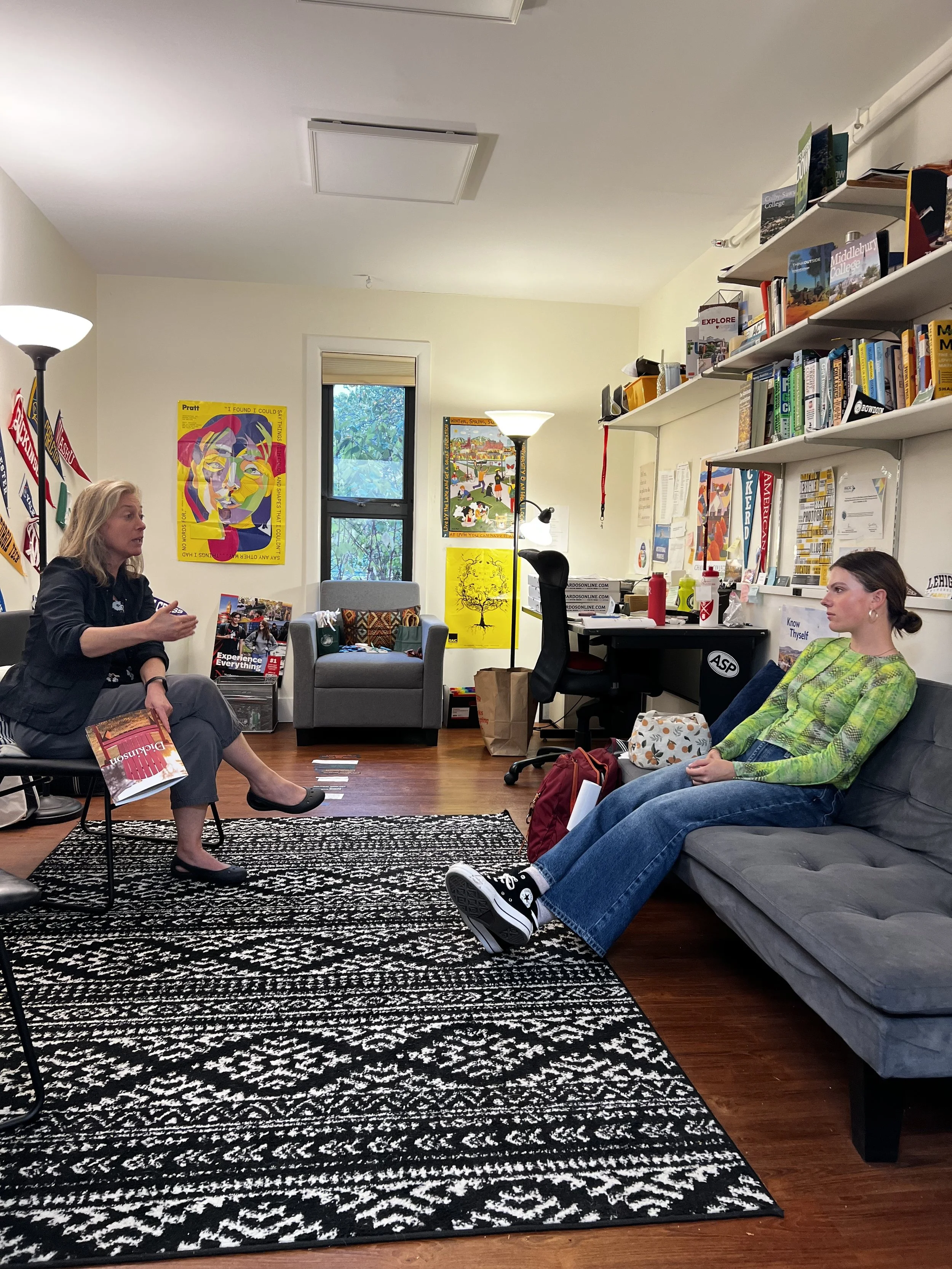 Two women in a conversation in a cozy office or classroom setting. One woman is sitting on a chair with a magazine in her lap, gesturing with her hand. The other woman is sitting on a sofa listening. The room has a black-and-white patterned rug, a gray armchair, bookshelves filled with books, and posters on the walls.