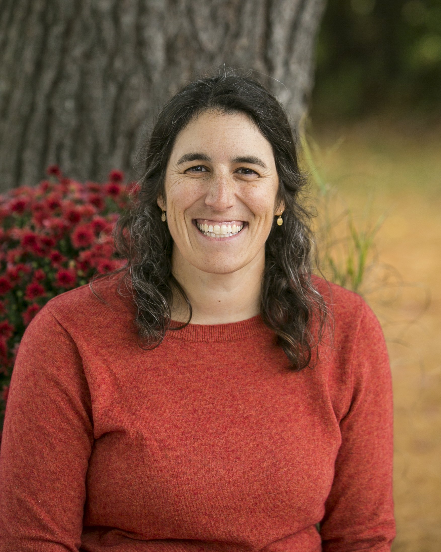 A smiling woman with dark curly hair, wearing a red sweater, outdoors near a tree and red flowers.