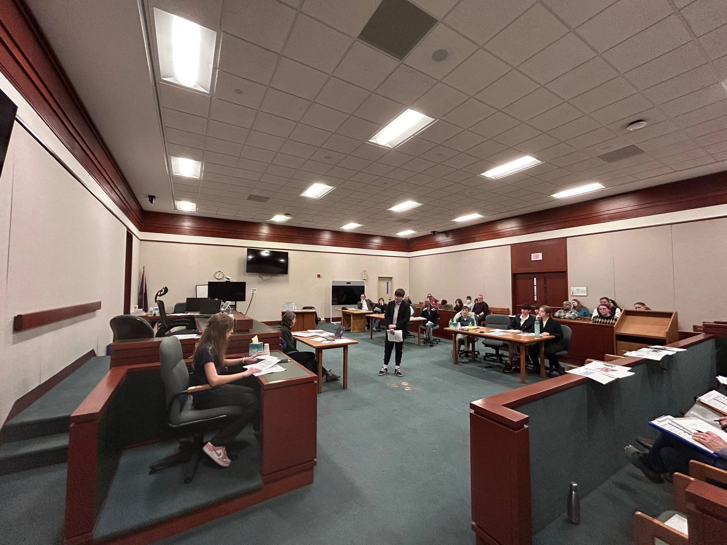 A courtroom with a judge sitting at the bench, several people seated at tables, and a person standing in the center with papers. The room has wooden furniture, a TV screen on the wall, and an exit door at the back.