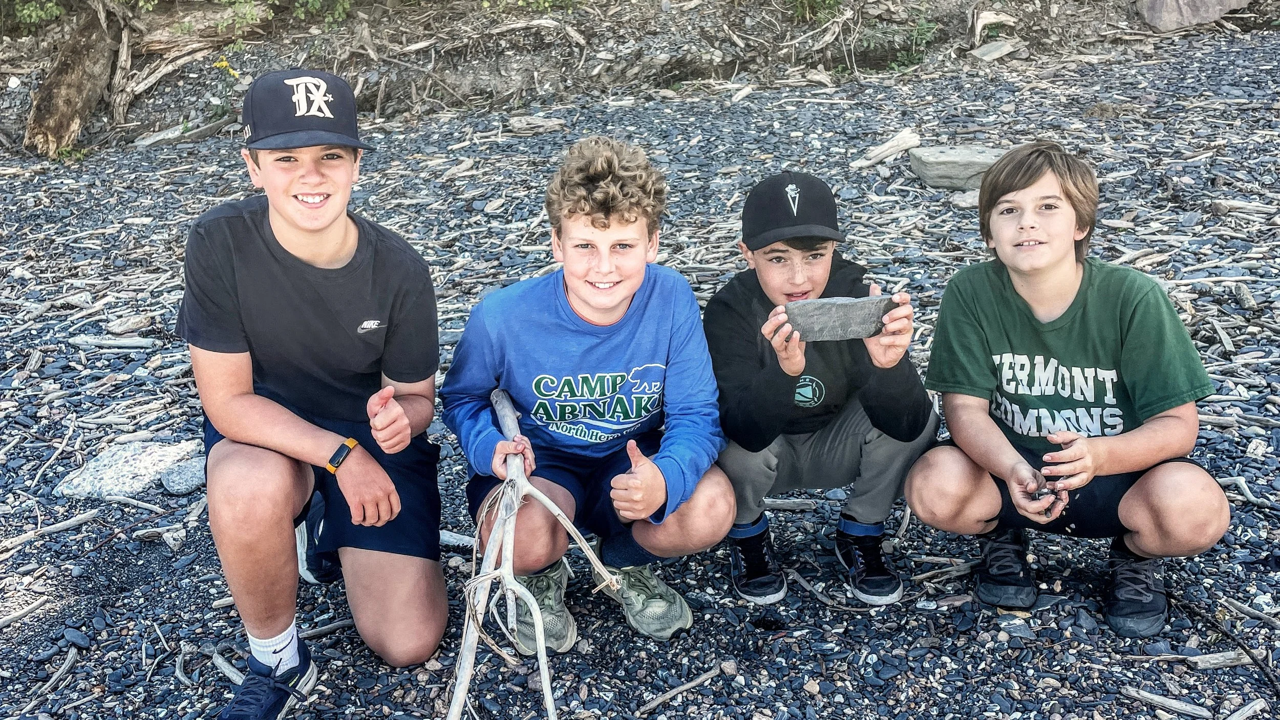 Four boys squatting on a rocky beach, smiling at the camera. One boy is holding a gray stone, another has a bunch of sticks, and a third is holding a smaller object. They are wearing casual clothes and hats, with a background of driftwood and small rocks.