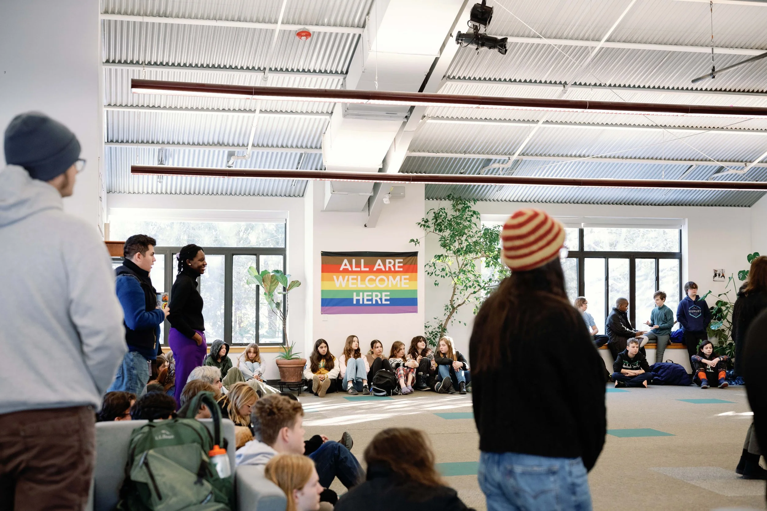 A diverse group of people gathered in an indoor space with large windows and potted plants. Some are sitting on the floor, others are standing, and a rainbow pride flag with the words "All Are Welcome Here" hangs on the wall.