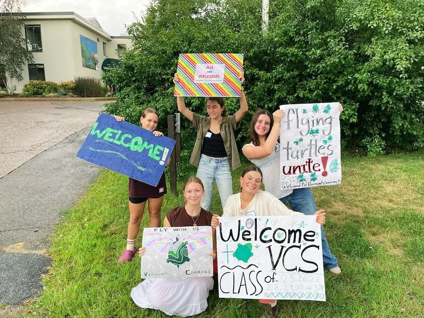 Group of five young women holding colorful welcome signs outdoors on a grassy area, trees, and a building in the background.