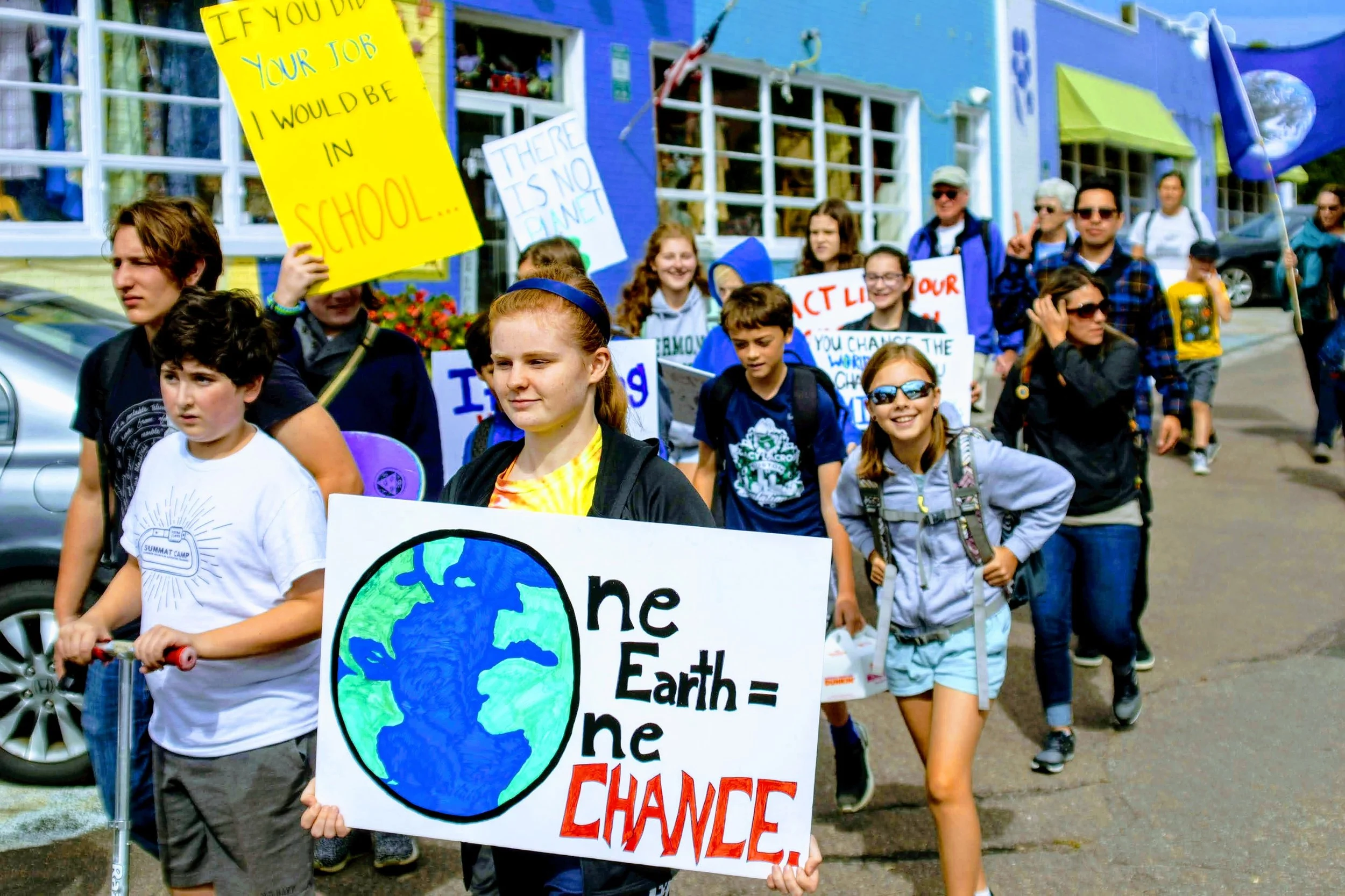 A group of children and adults participate in a climate protest march on a city street. The children are holding handmade signs about climate change and environmental awareness, including one depicting the Earth with the message "the Earth = one CHAN