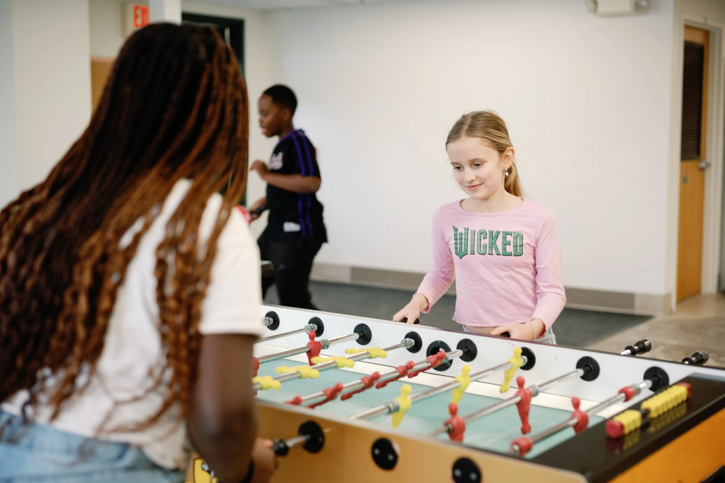 Two girls playing foosball in a recreational room, with a boy in the background.