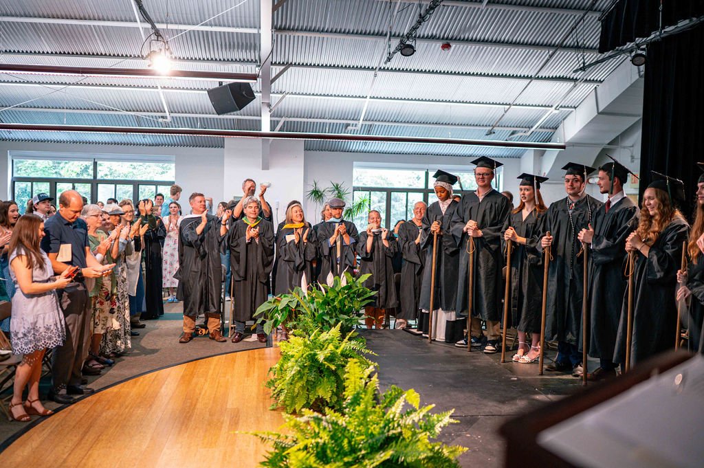 Group of graduates in black caps and gowns holding staffs, standing on stage at graduation ceremony, with audience clapping and taking photos, indoor setting with ceiling lights and large windows.