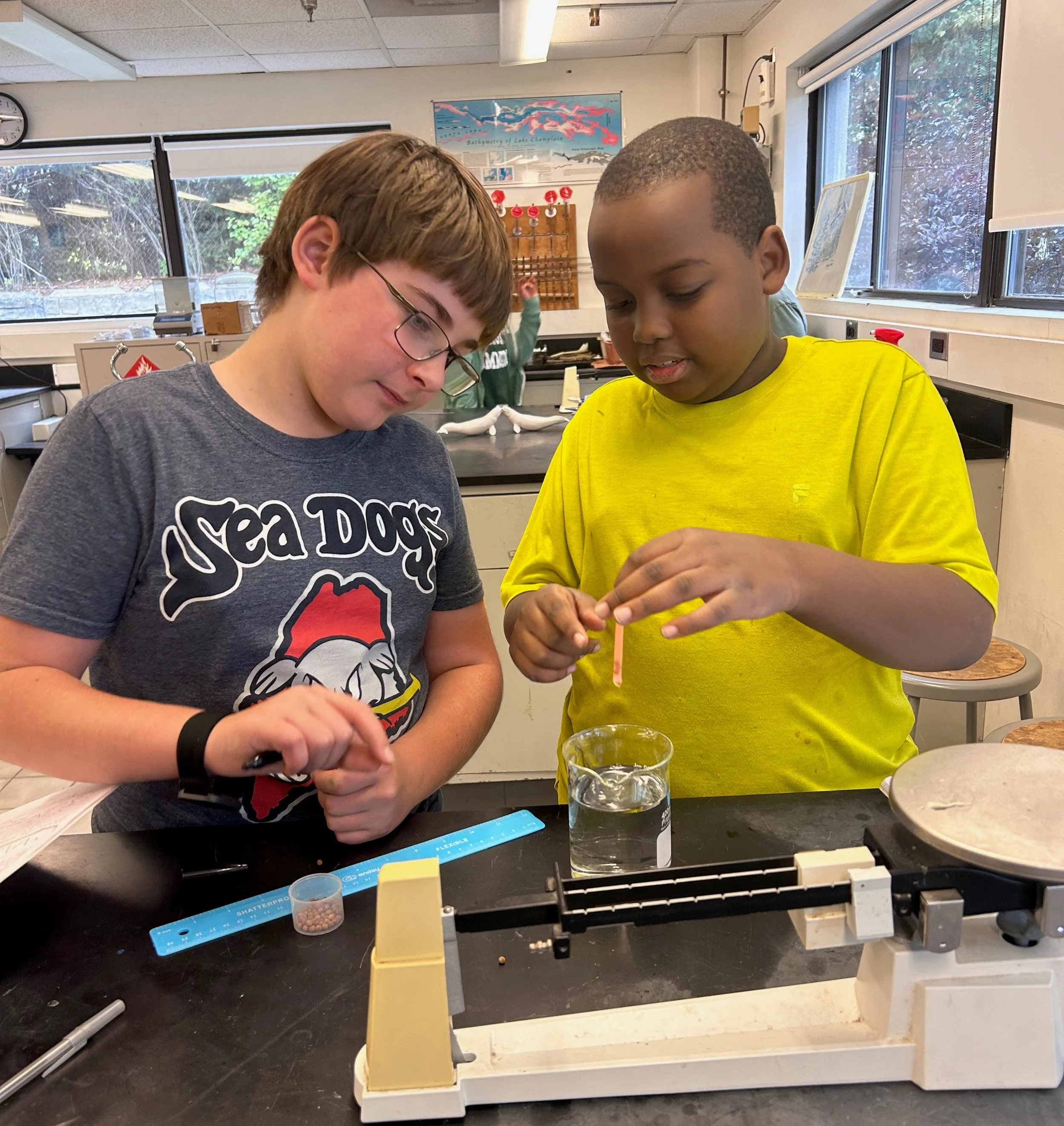 Two boys conducting a science experiment in a classroom; one wearing a gray 'Sea Dogs' shirt and glasses, the other in a yellow shirt, holding a test tube over a beaker of water.