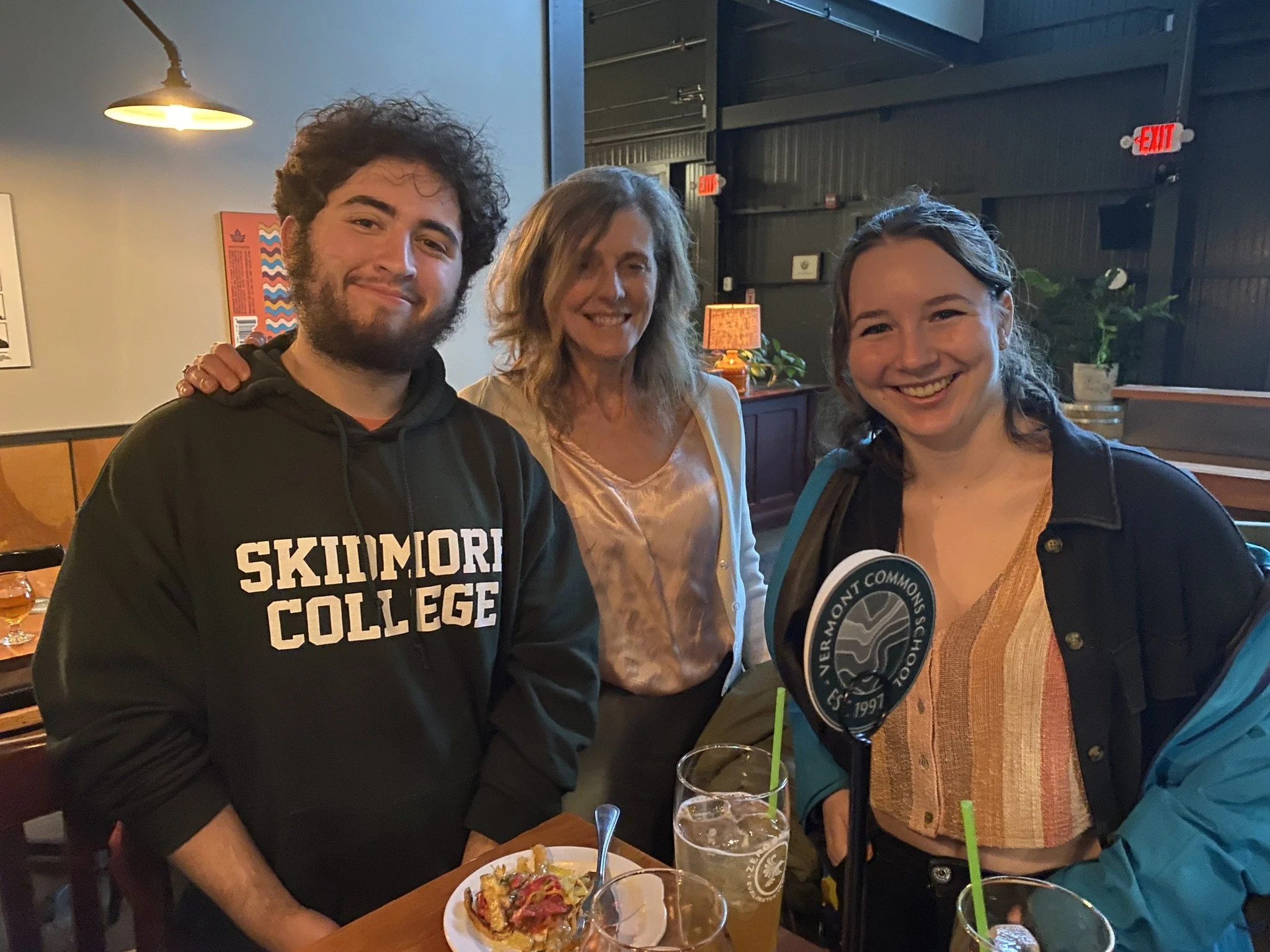Three people smiling at a table in a restaurant, with two glasses of drinks in front of them. The person on the left is a young man with curly hair and a beard, wearing a black hoodie that says 'Skimmort College.' The woman in the middle has shoulder-length blonde hair and is wearing a light-colored blouse. The person on the right is a young woman with long hair, wearing a black jacket over an orange and beige striped shirt, holding a sign that reads 'Vermont Common School'.