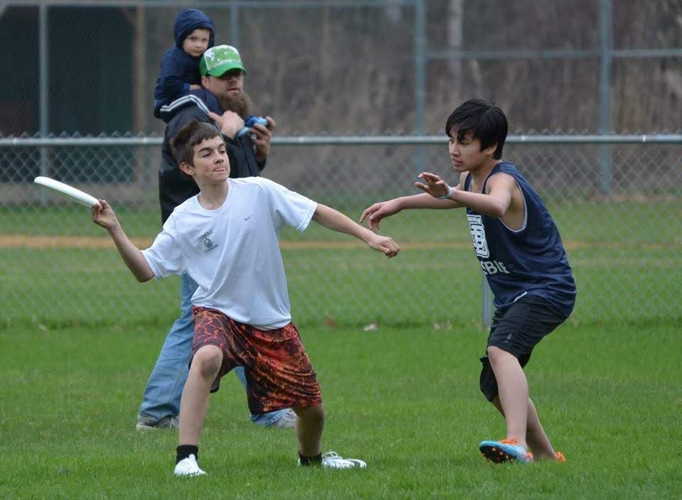 Two boys play catch with a frisbee on a grassy field, with a chain-link fence in the background. An adult man and a young child watch nearby.