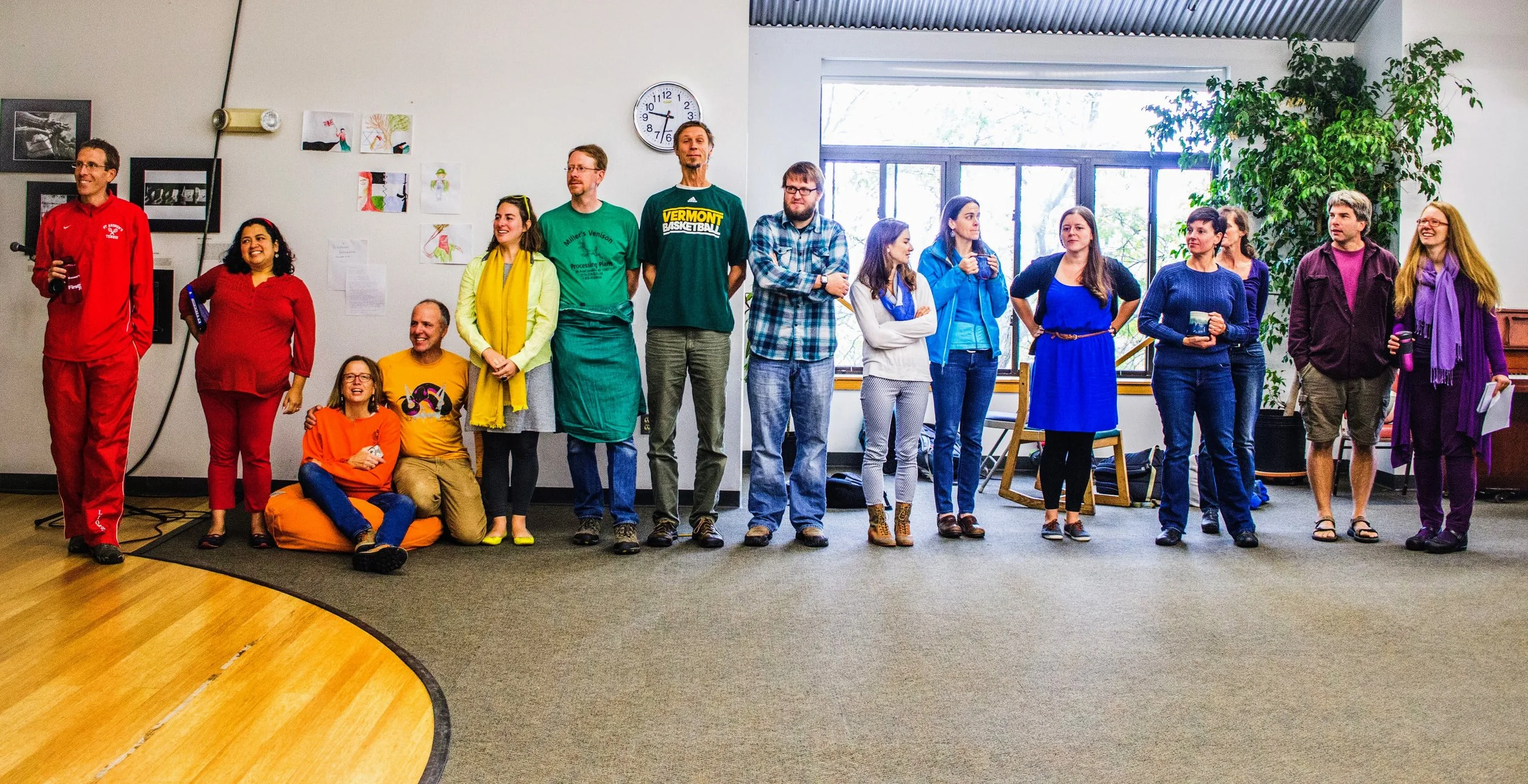 Group of diverse people standing and sitting in a room with art on the wall, large window, and indoor plants, engaging in a gathering or meeting.