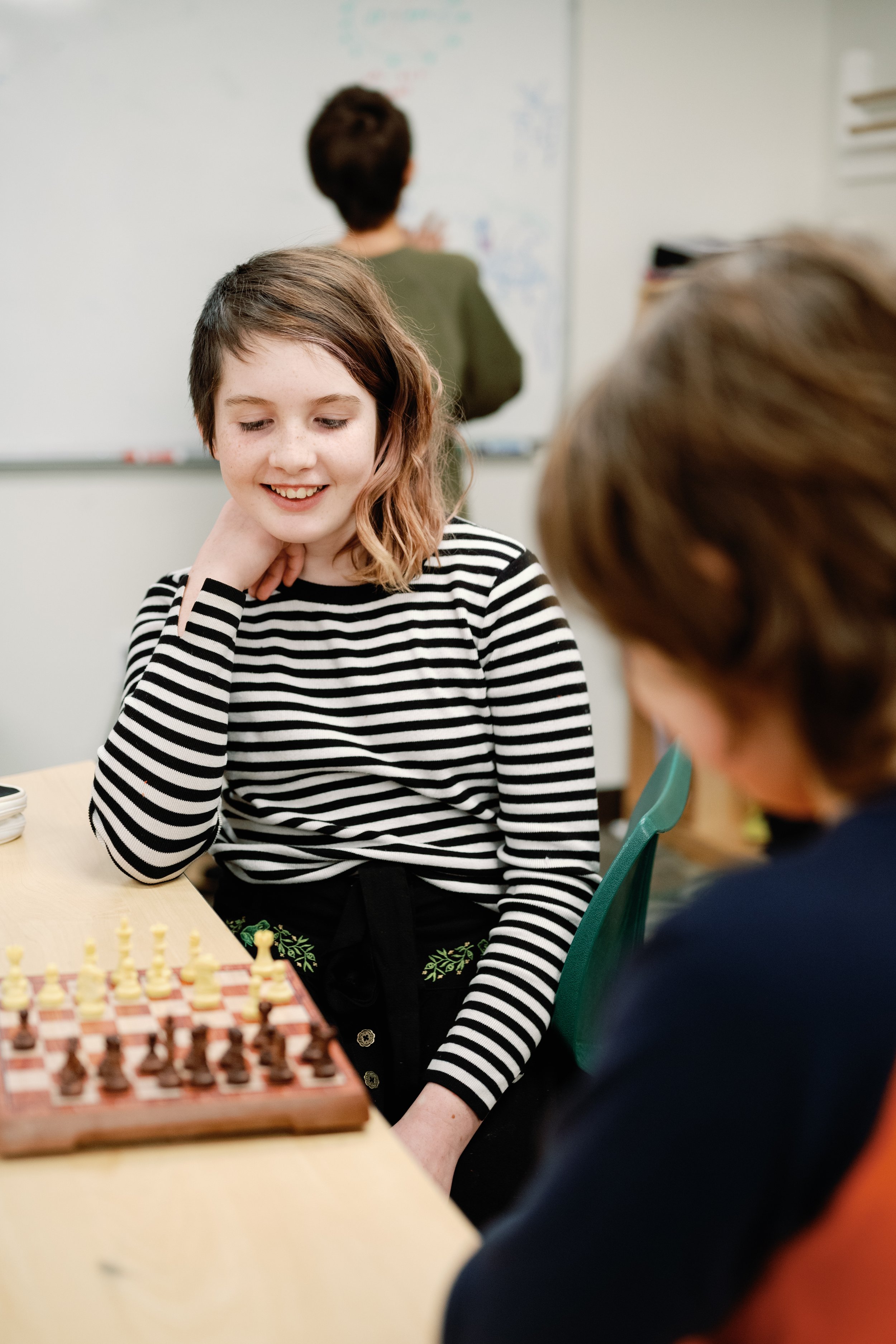 A girl with shoulder-length brown hair and light skin, wearing a black and white striped shirt, sitting at a table and smiling while playing chess with a boy with light skin and brown hair.