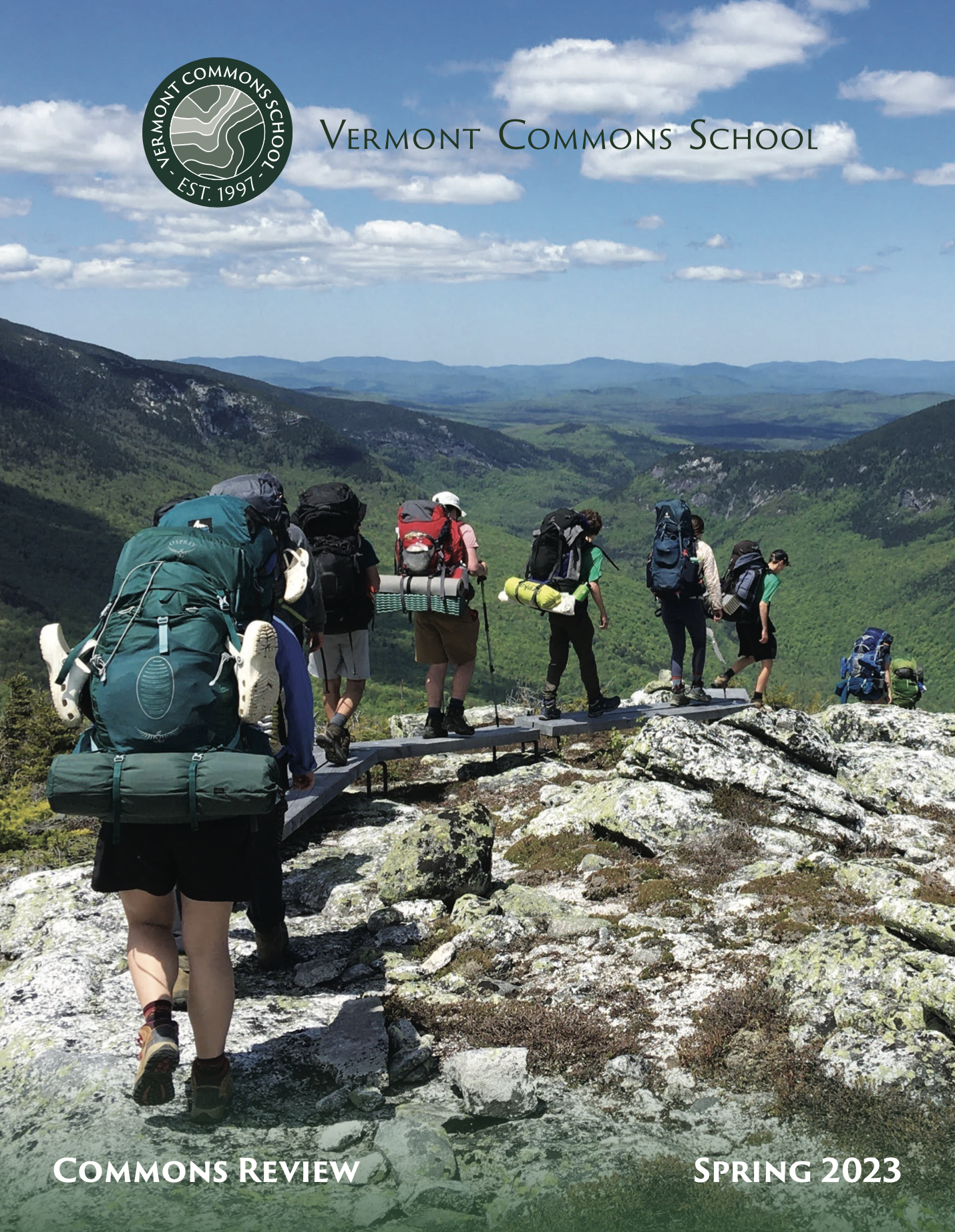 Group of hikers walking across a small bridge in a mountainous landscape, with green hills and valleys under a blue sky with scattered clouds.